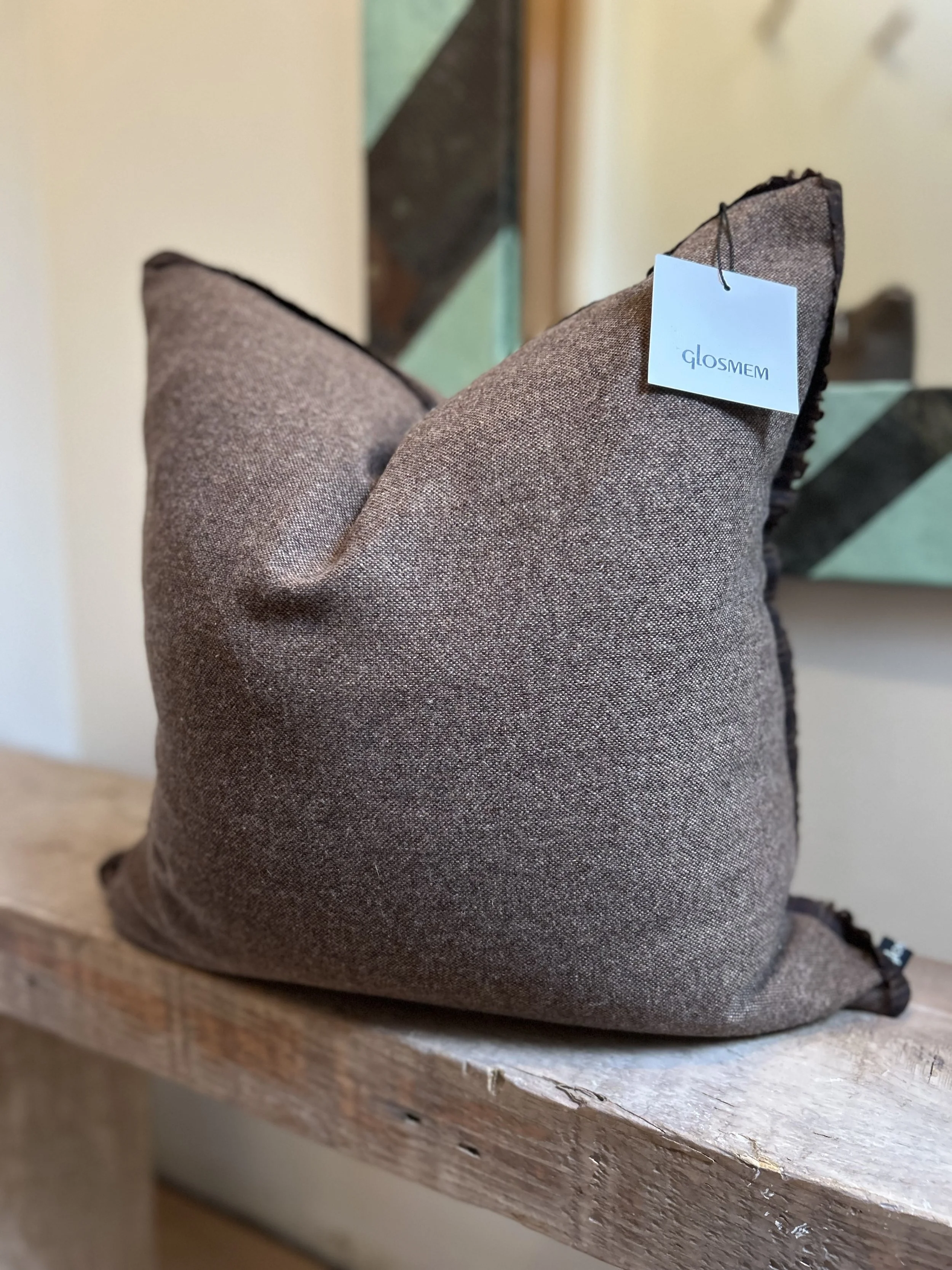 A brown fabric pillow with black edges, placed on a wooden surface. The pillow has a white tag attached to it that reads "GLOSME".