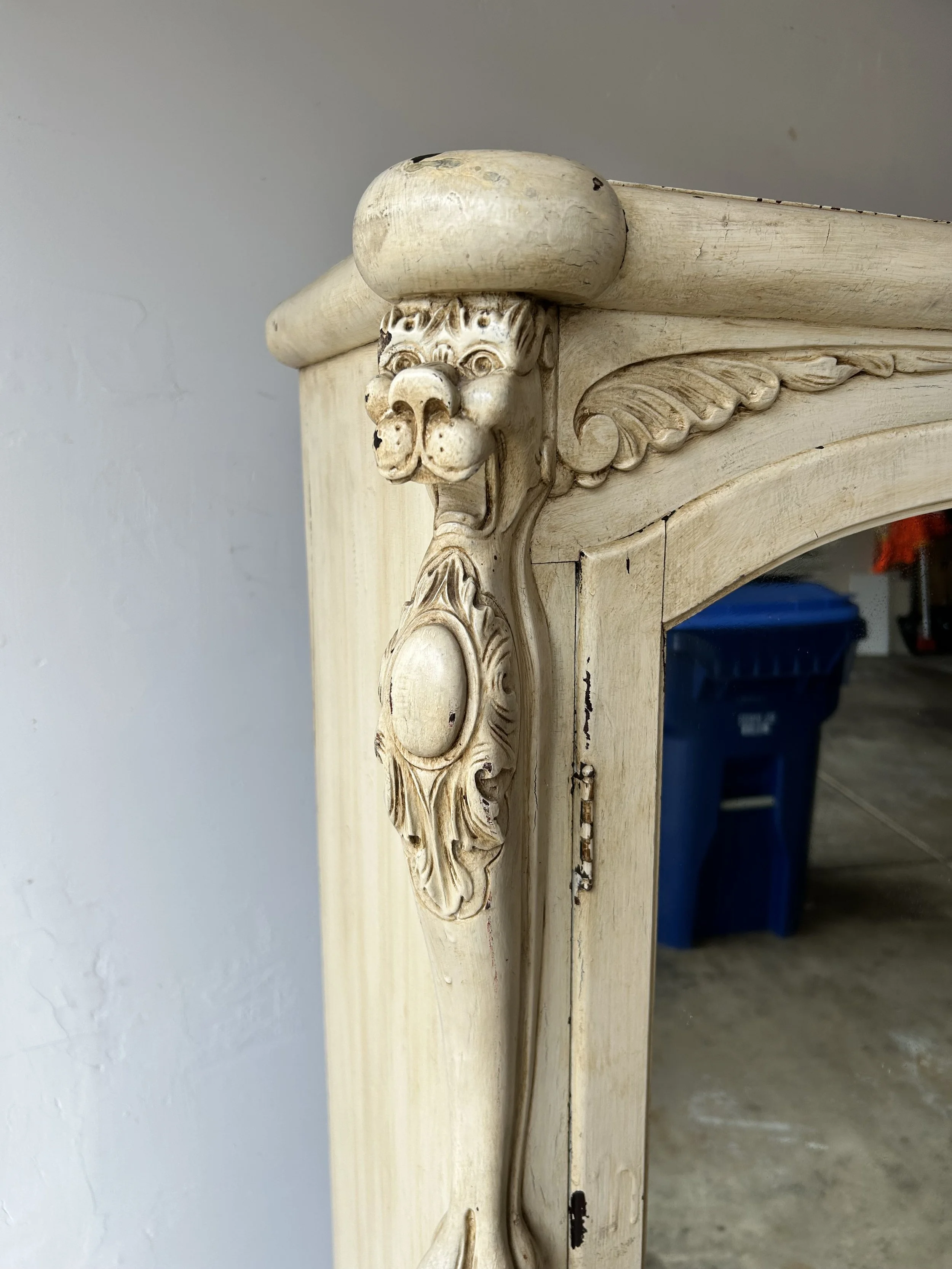 Close-up of an ornate antique cream-colored wooden furniture piece with a carved lion's head detail at the top corner, showing signs of wear.