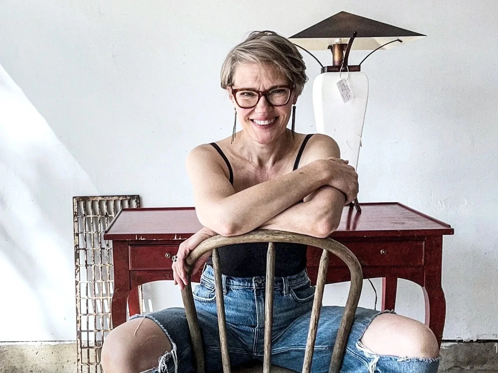 A smiling woman with short gray hair and glasses, wearing a black tank top and ripped denim shorts, is sitting on the floor in front of a wooden chair, leaning on the back of the chair with her arms crossed over it. Behind her, there is a red wooden desk and a modern table lamp with a white base and black shade, against a plain white wall.