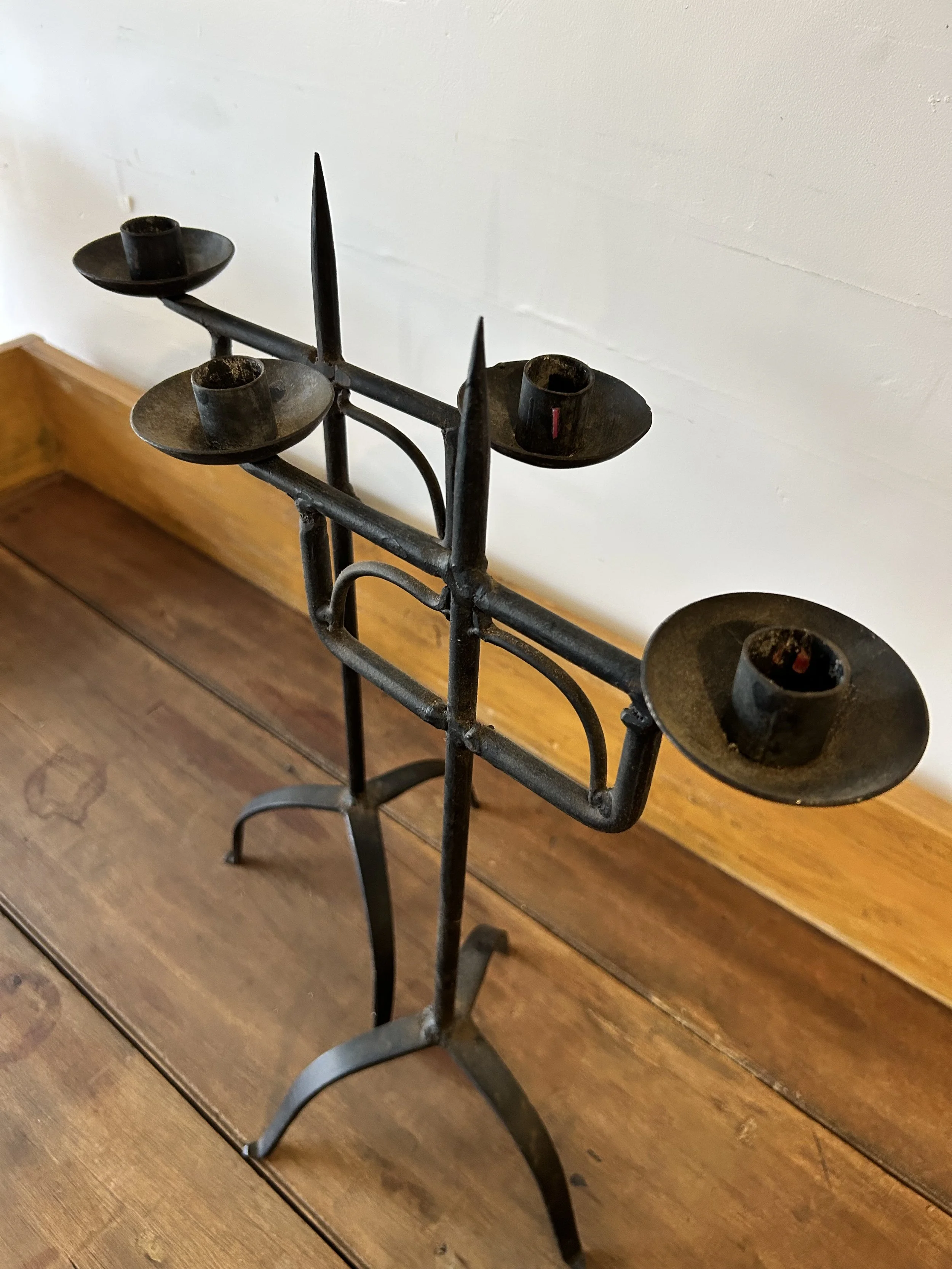 A vintage black metal candle holder with six circular bases for candles, standing on a wooden surface against a white wall.