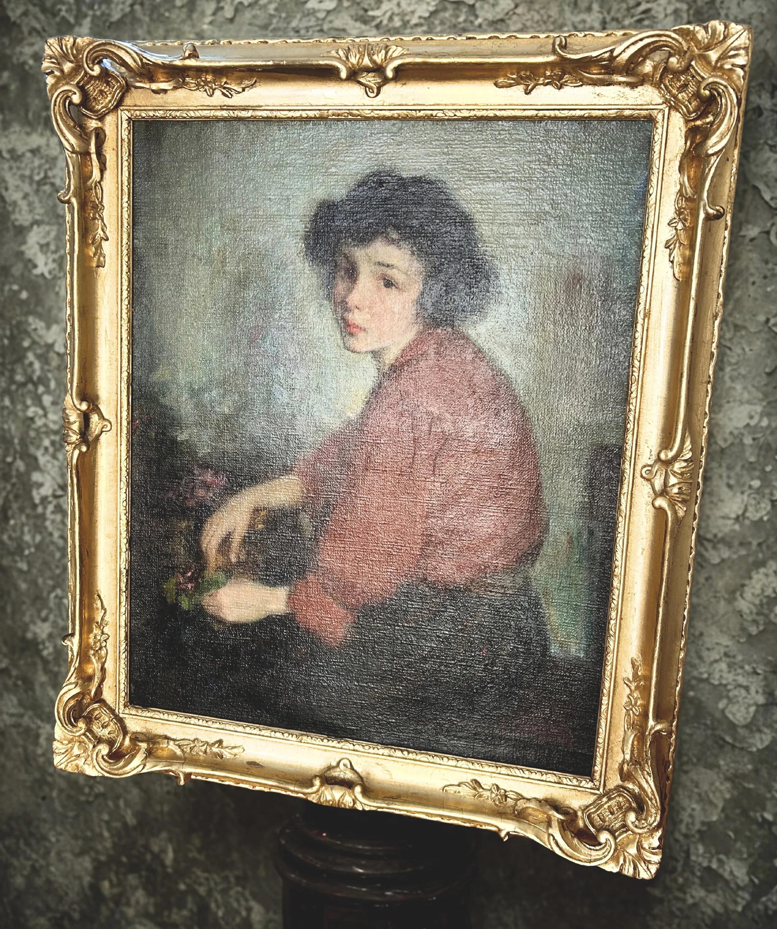 A framed oil painting of a woman with short curly black hair, wearing a pinkish-red top, sitting and looking to the side while holding a small plant or flower in her hand. The background of the painting is muted and textured.
