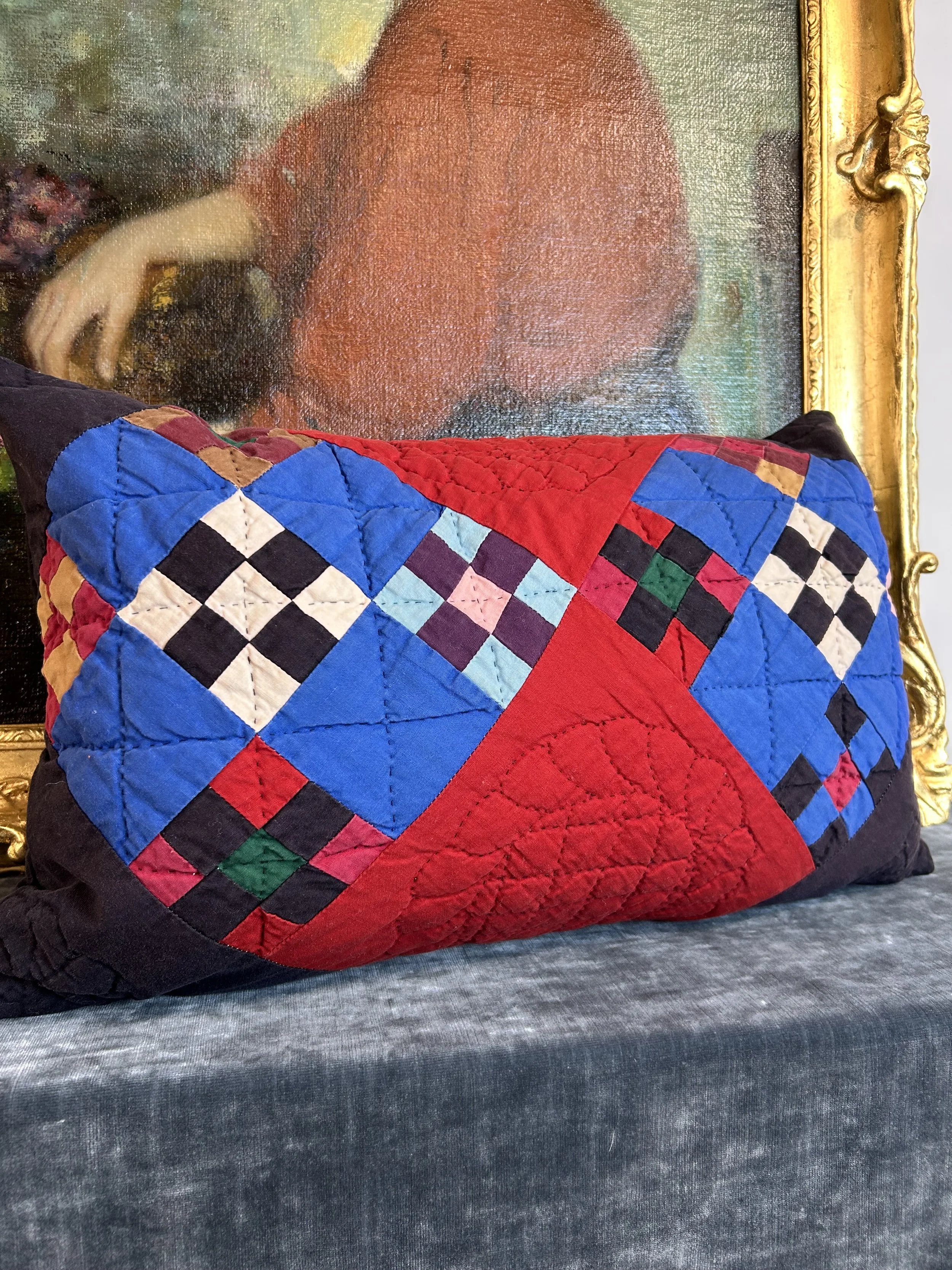 Colorful quilted pillow with a geometric pattern featuring red, blue, black, white, and pink fabric, placed on a dark fabric surface in front of a framed classical painting.