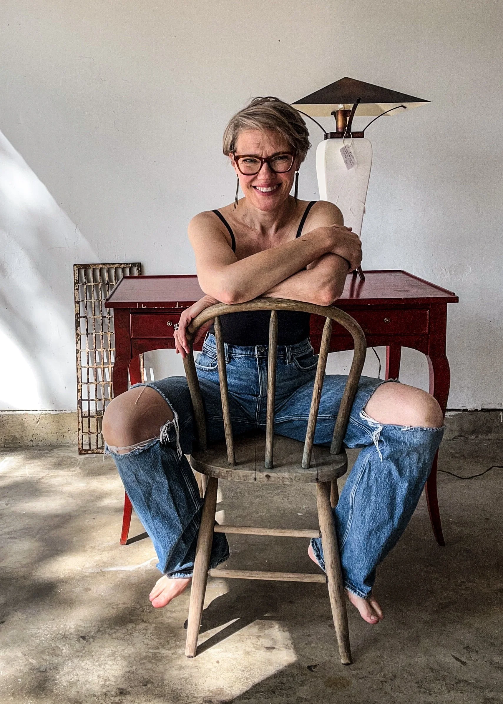 A woman with short blonde hair, wearing glasses, a black tank top, and ripped jeans, sitting on a wooden chair with her legs spread wide. She is smiling and resting her arms on the back of the chair. In front of her, there is a red wooden table and a white lamp on top of it. The setting appears to be indoors with concrete flooring and a white wall.