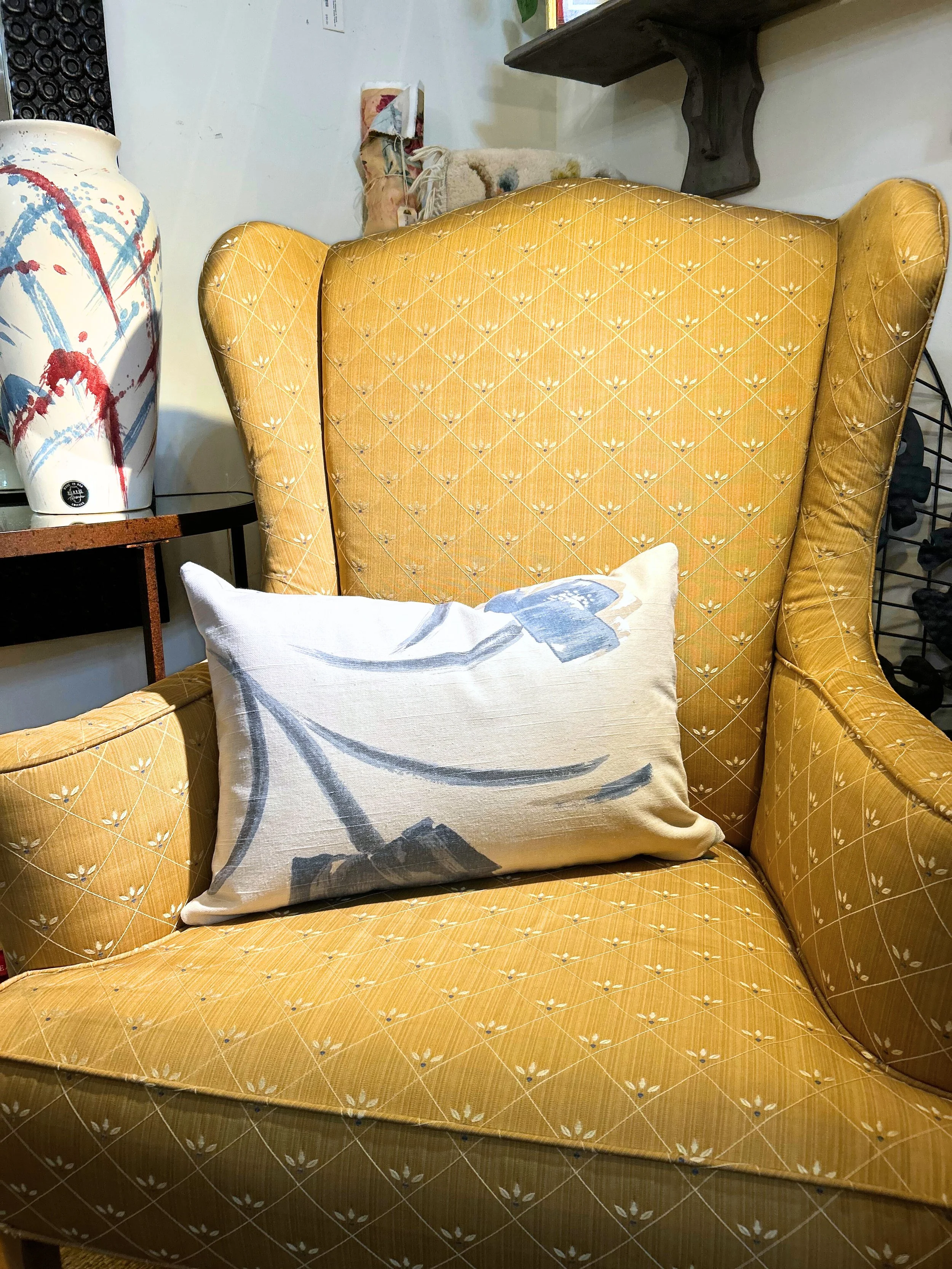 A yellow upholstered armchair with a high, rounded back and armrests, decorated with a diamond pattern and small floral details, with a light-colored cushion featuring a floral design resting on it, in an indoor setting with decorative items and furn