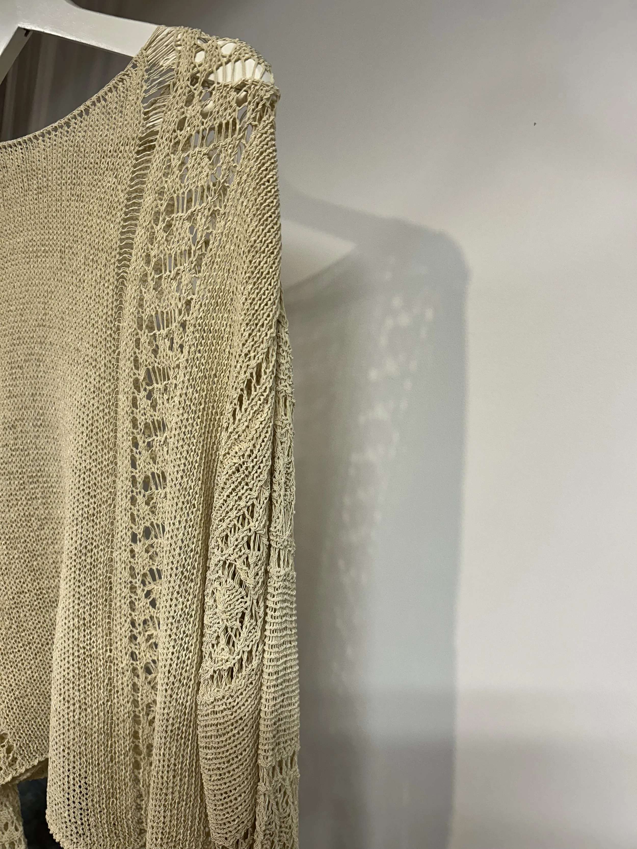 A beige, intricately crocheted or knitted garment hanging on a hanger, casting a detailed shadow on a white wall.