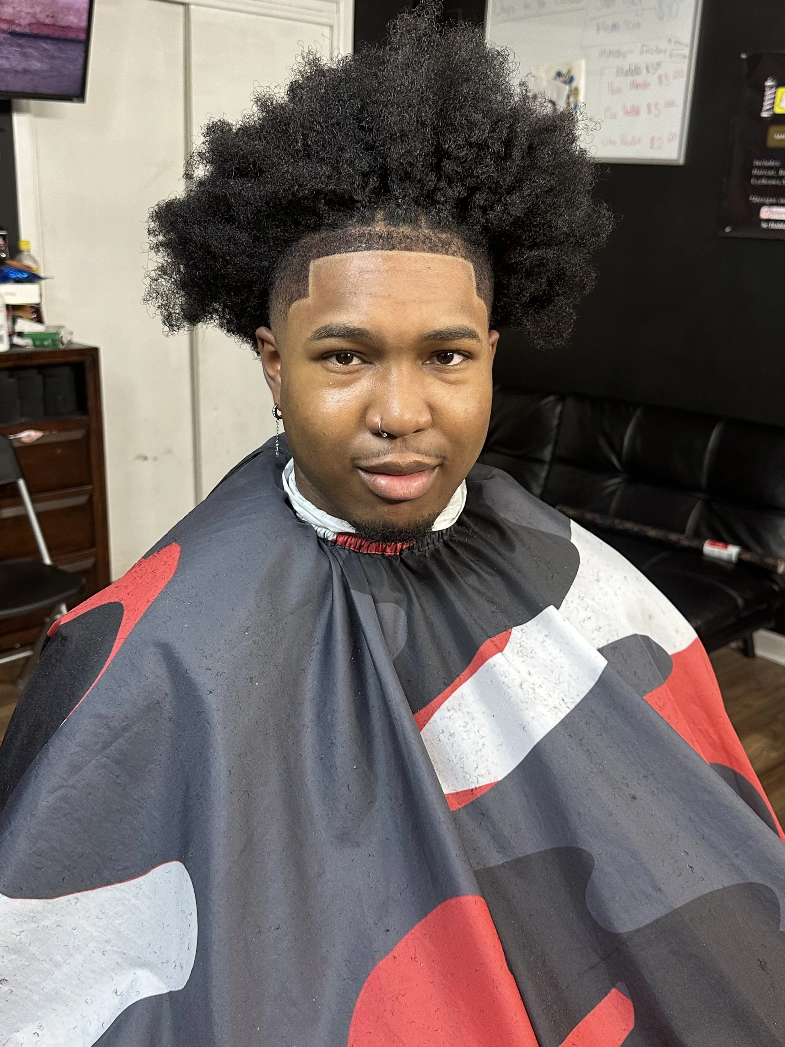Young man with a fresh fade haircut sitting in a barber's chair, wearing a black, red, white, and gray barber cape in a barbershop.