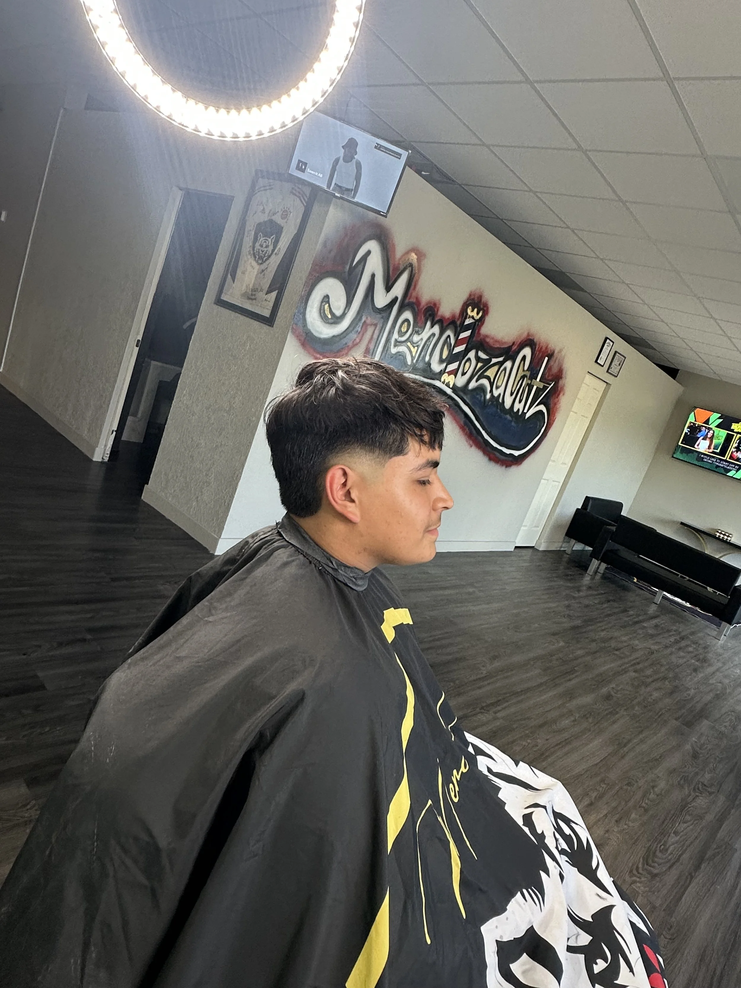 Man getting a haircut in a salon with a wall hanging graffiti-style sign that says 'Merzo'z Cutz'.