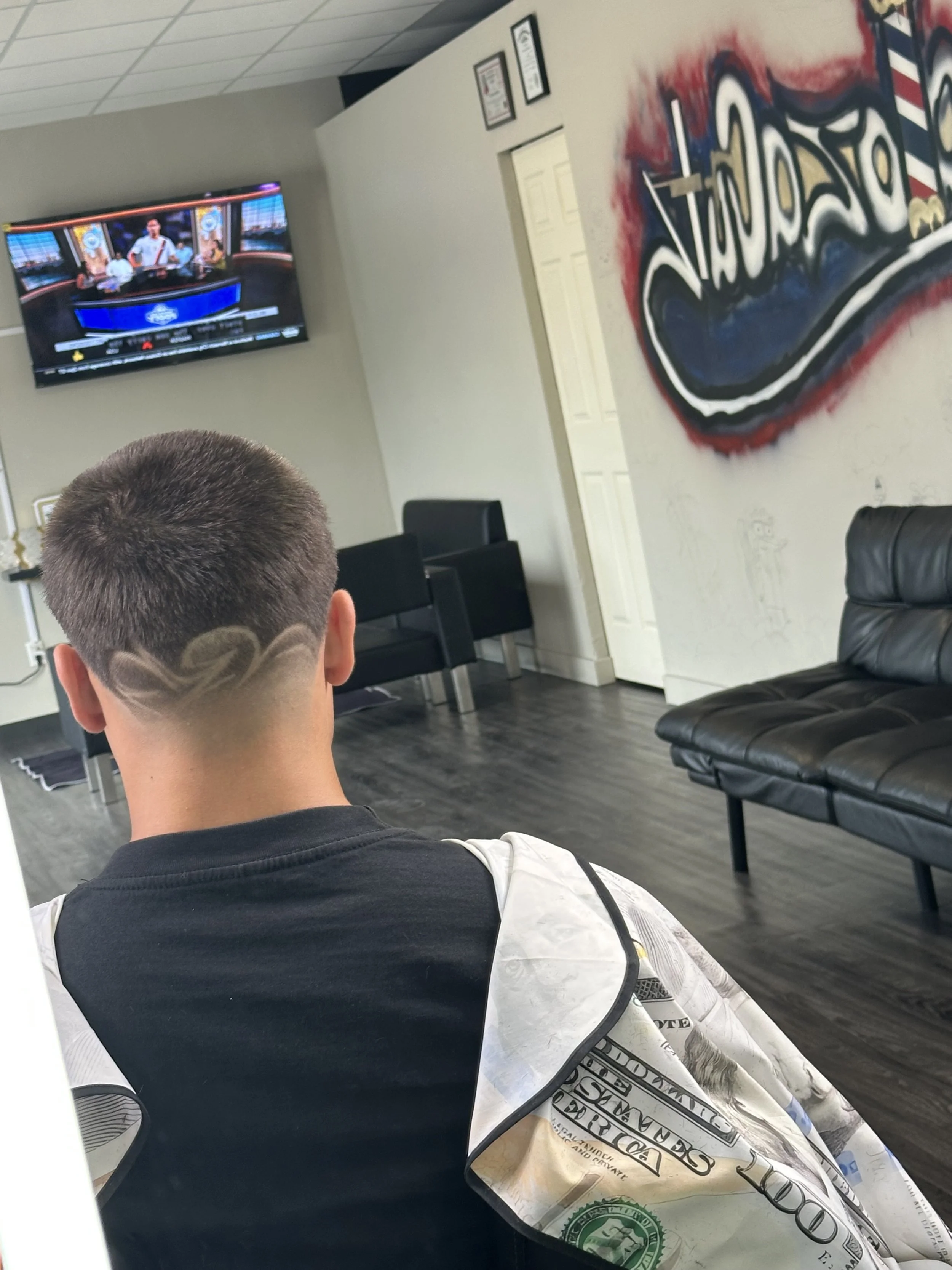 A person sitting in a barbershop with a haircut featuring a wave design shaved into the back of their head, wearing a cape made from a hundred-dollar bill.