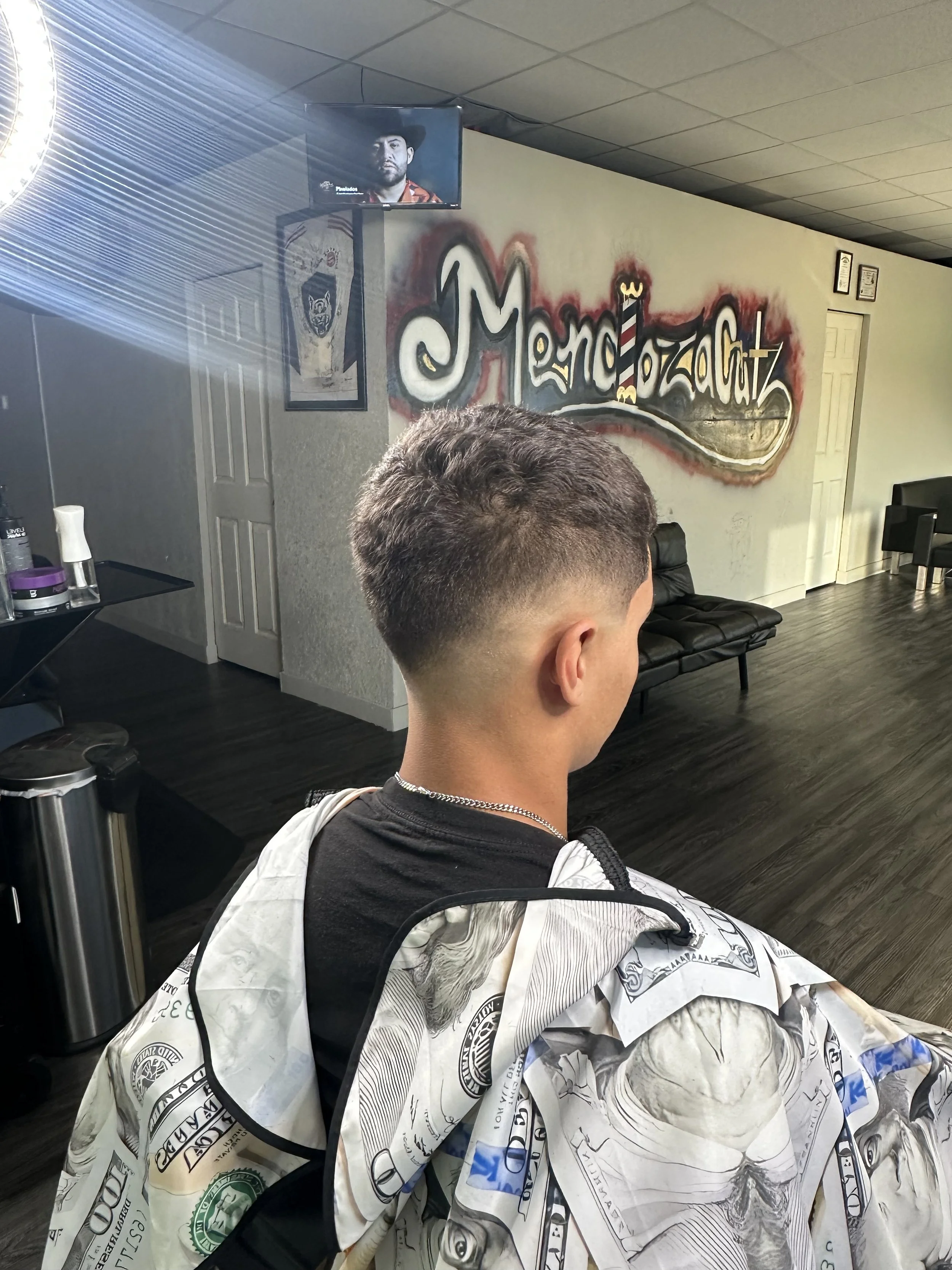 A young man with a short, fade hairstyle sitting in a hair salon, with a cape made of hundred-dollar bills around his shoulders. The salon features a graffiti-style mural on the wall with the words "MonkeyzazGutz" and a TV mounted on the wall display