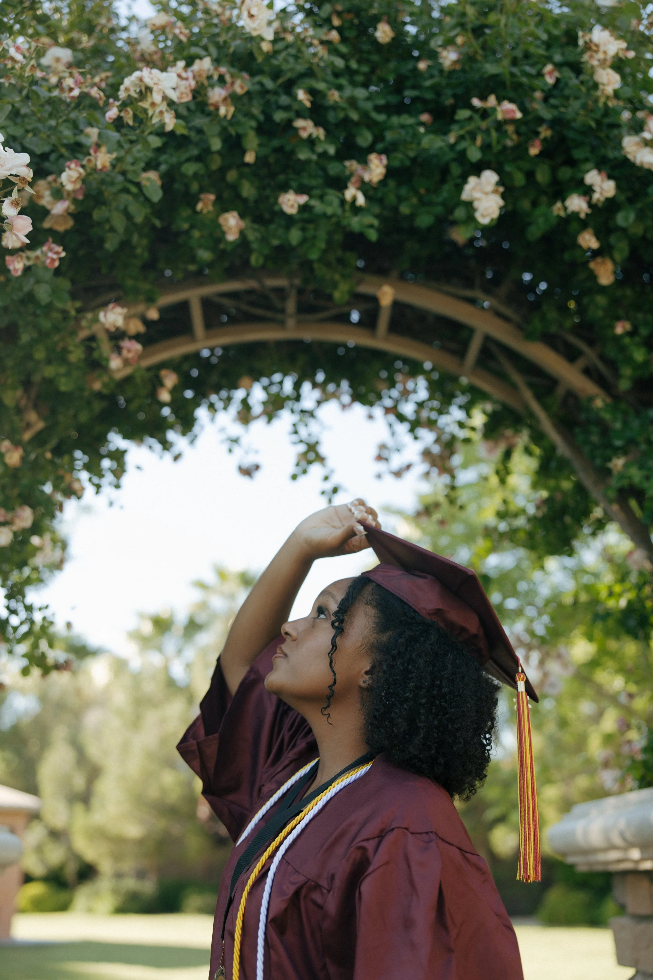 A young woman in a maroon graduation gown and cap adjusts her cap while standing outdoors under a flowering archway.