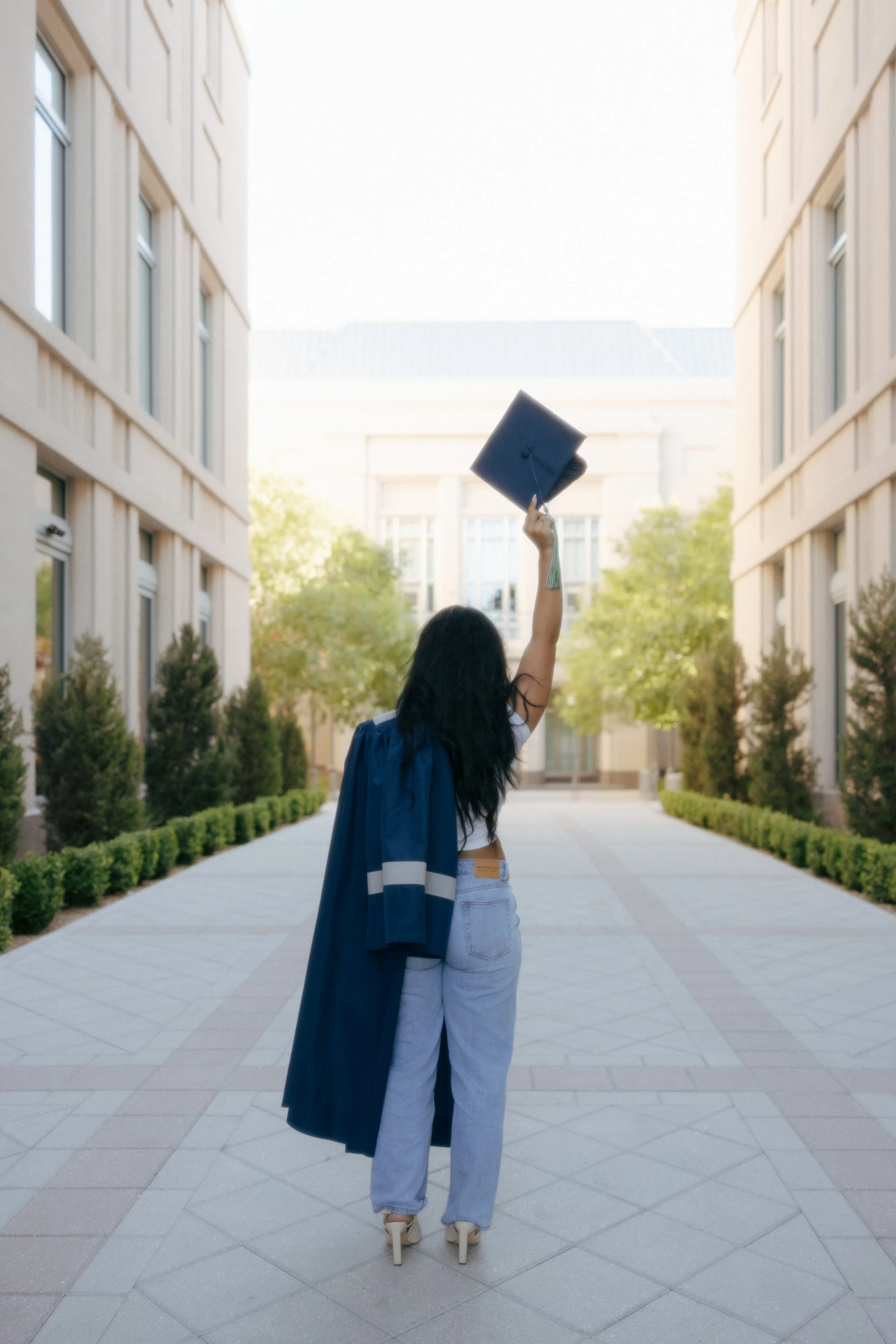 A woman in casual jeans and heels, with a graduation gown draped over her shoulders, holding a graduation cap high in the air, standing outdoors on a paved path between two modern buildings with trees and bushes.
