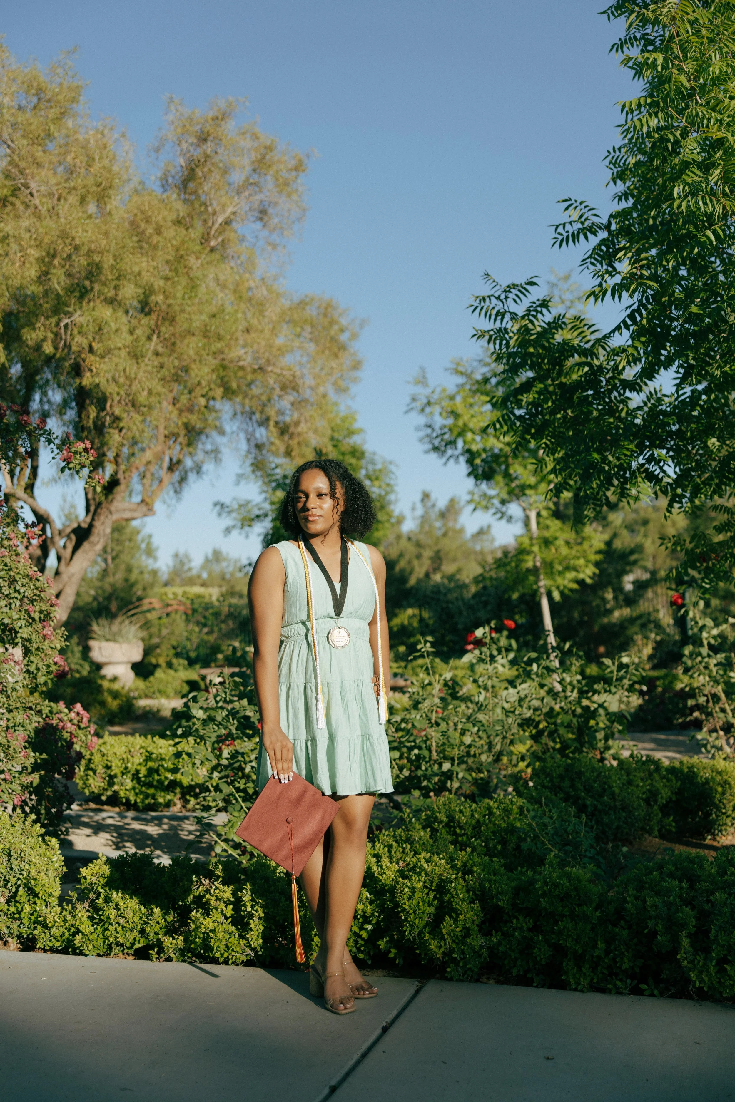 A woman in a light green dress holding a brown diploma folder standing on a sidewalk in a lush garden with trees and shrubs under a clear blue sky.