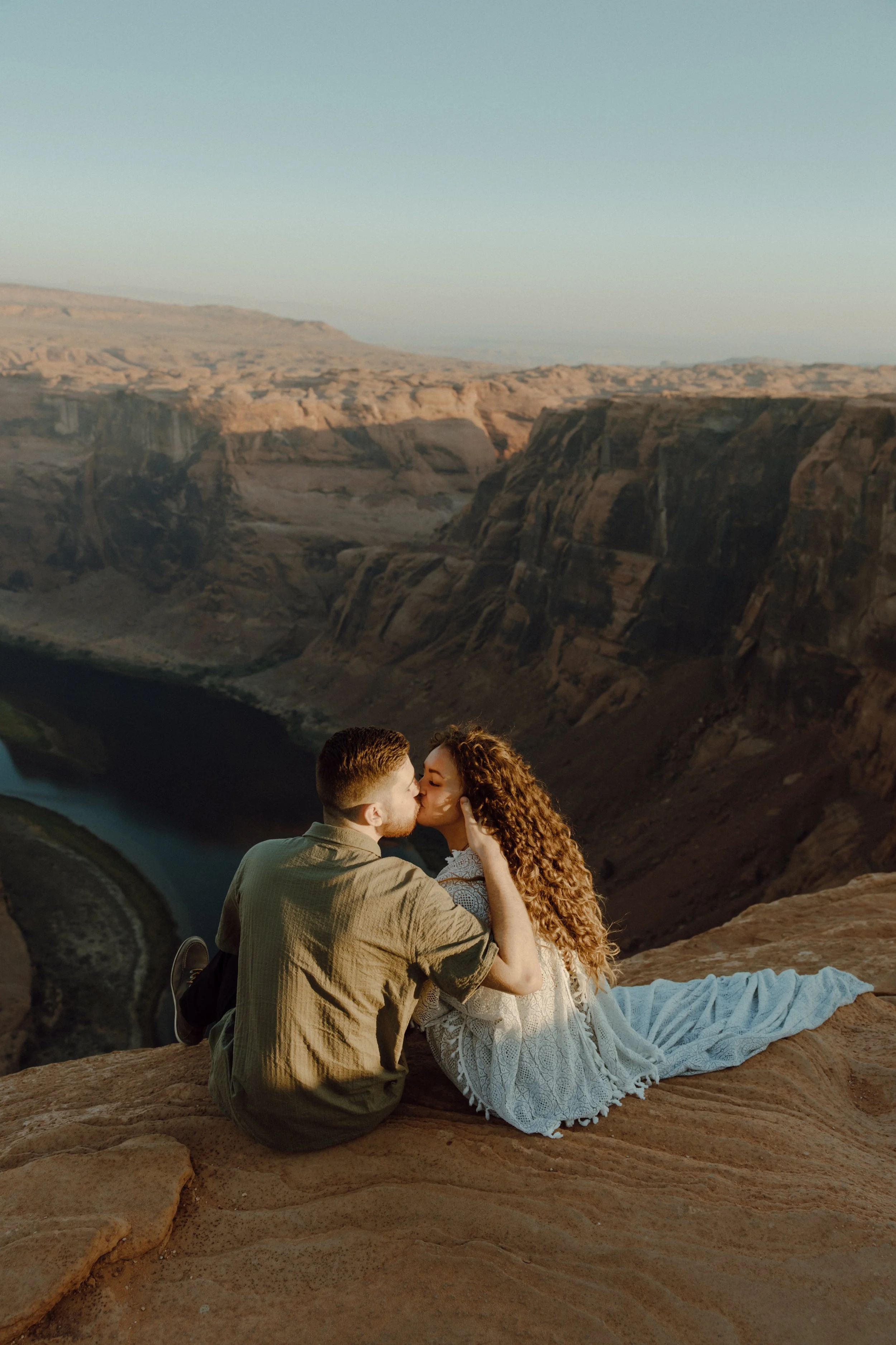 A couple sharing a kiss while sitting on a rocky ledge overlooking a canyon with a river below at sunset.