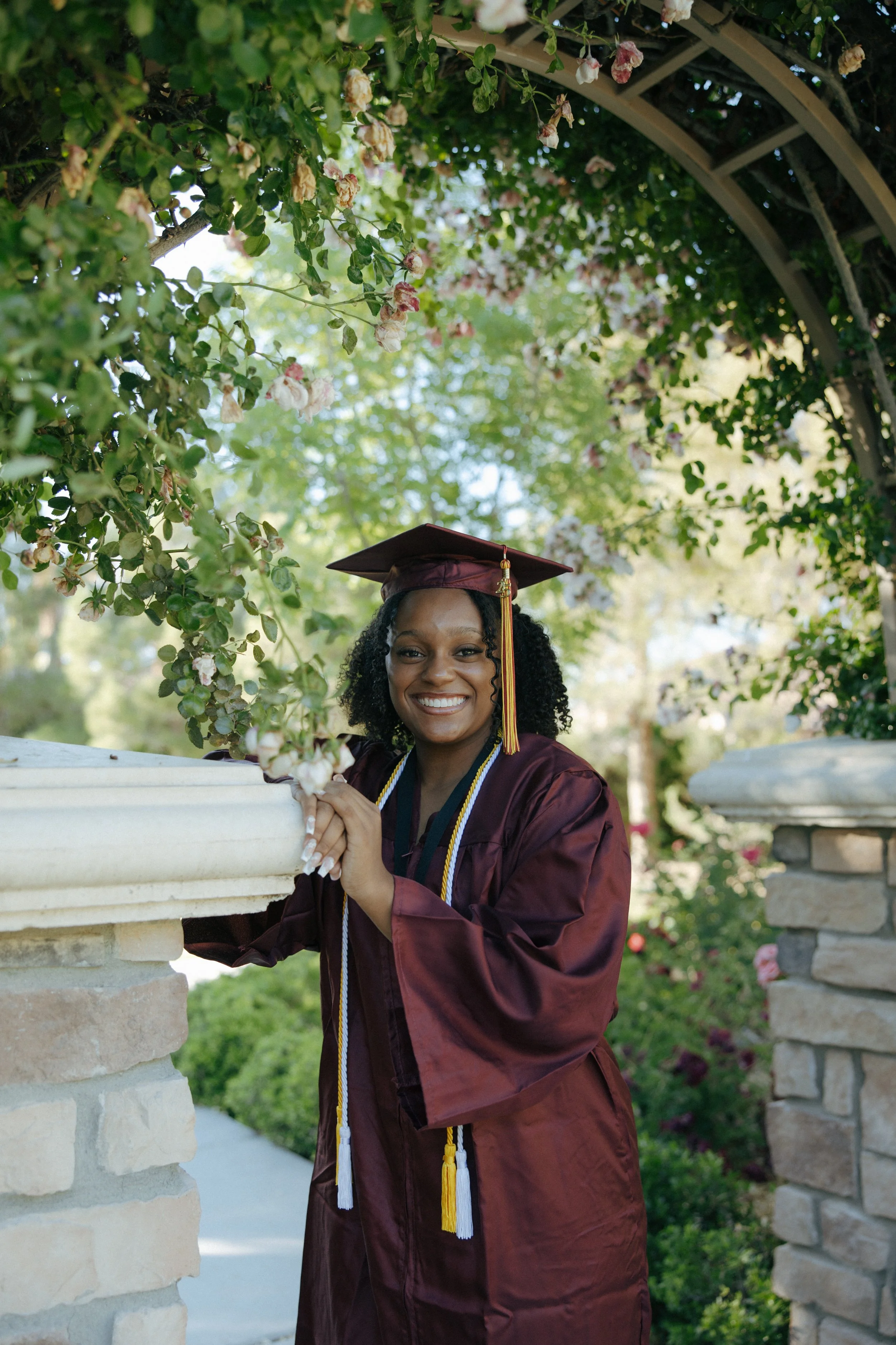 A woman in a maroon graduation gown and cap stands outdoors under a flowering archway, smiling and holding onto a stone and wooden garden structure.