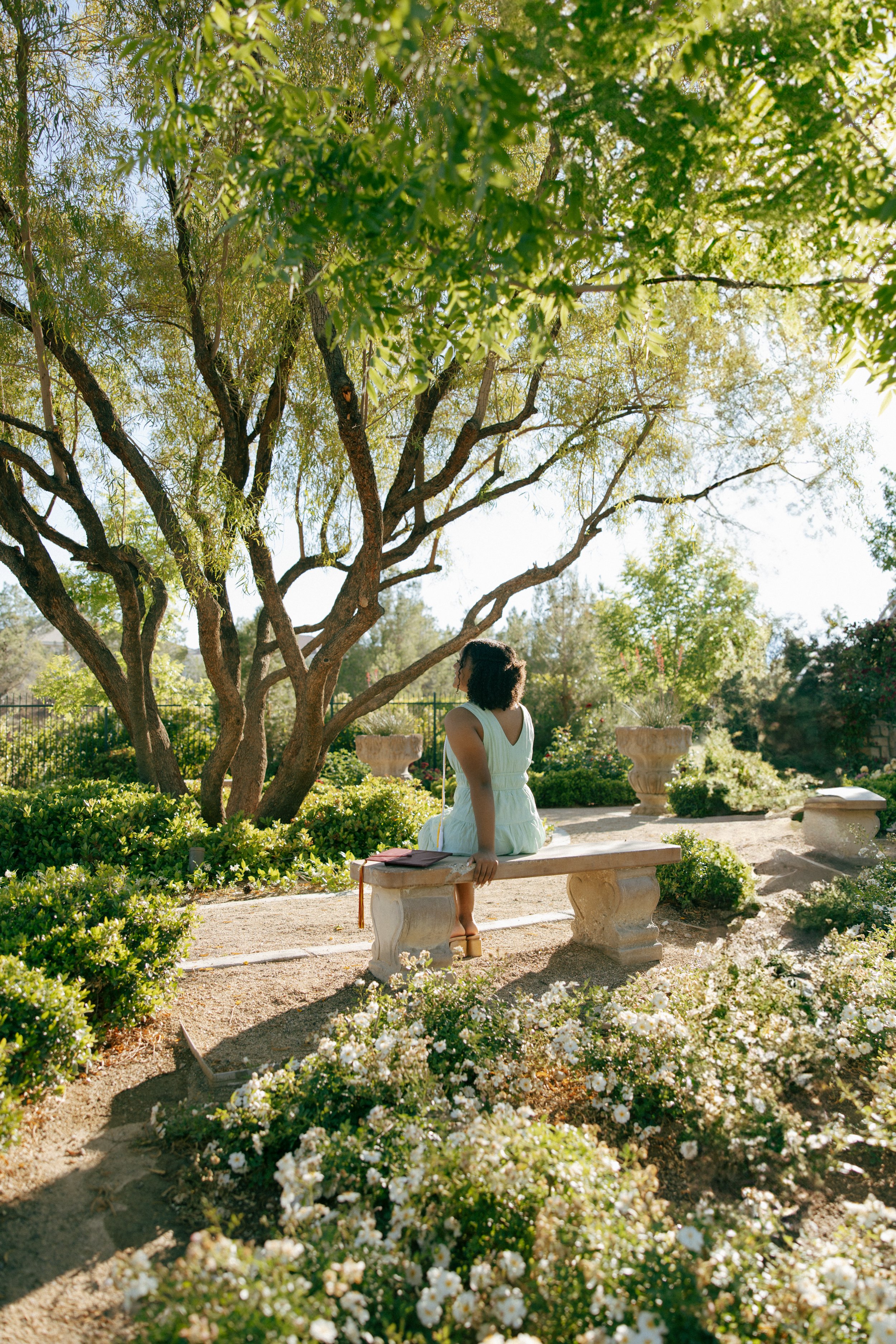 A woman in a light dress sitting on a stone bench in a lush garden, with a large tree and flowering bushes nearby, enjoying the sunlight.