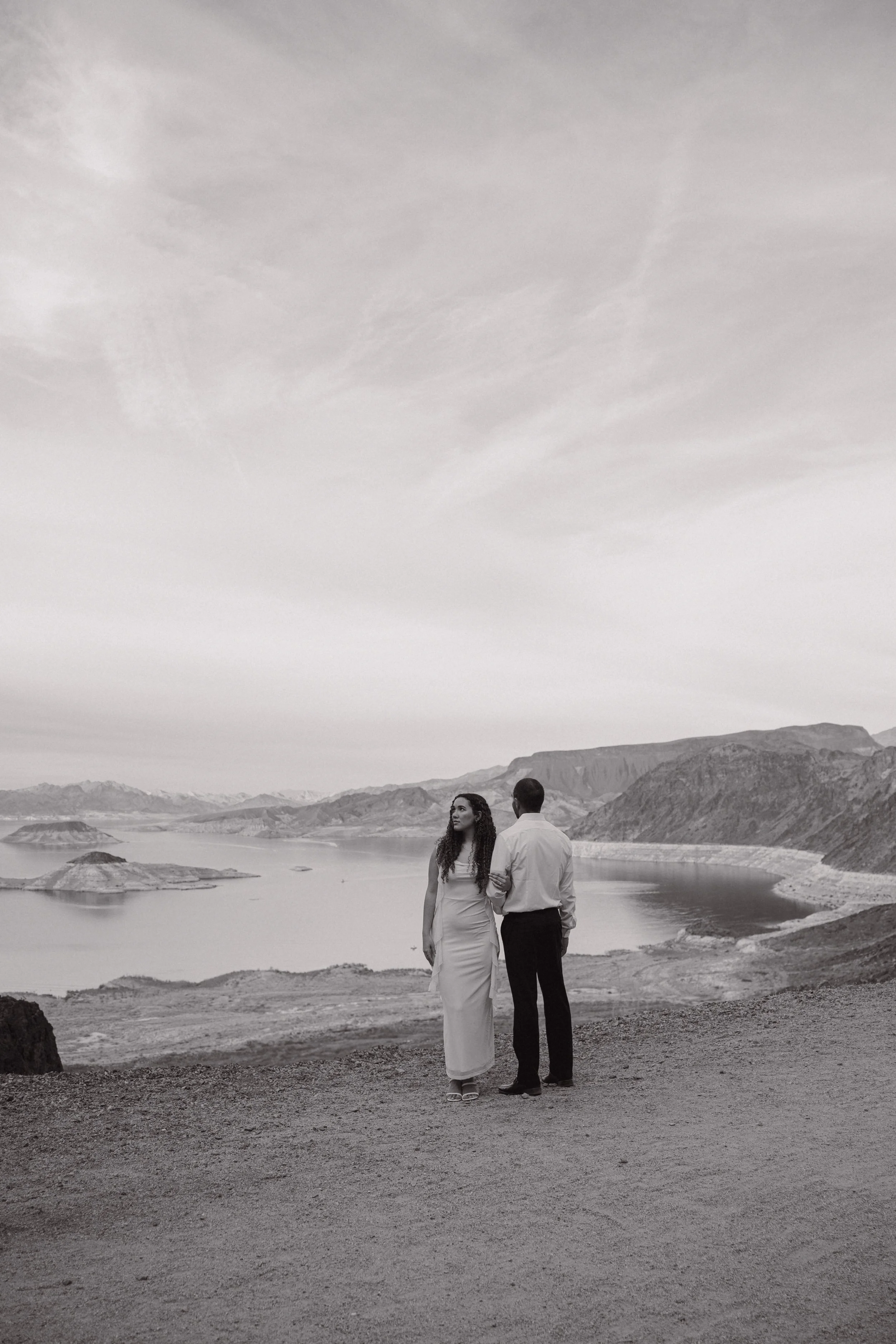 A black and white photo of a couple standing on a dirt path overlooking a large body of water, mountains, and sky.