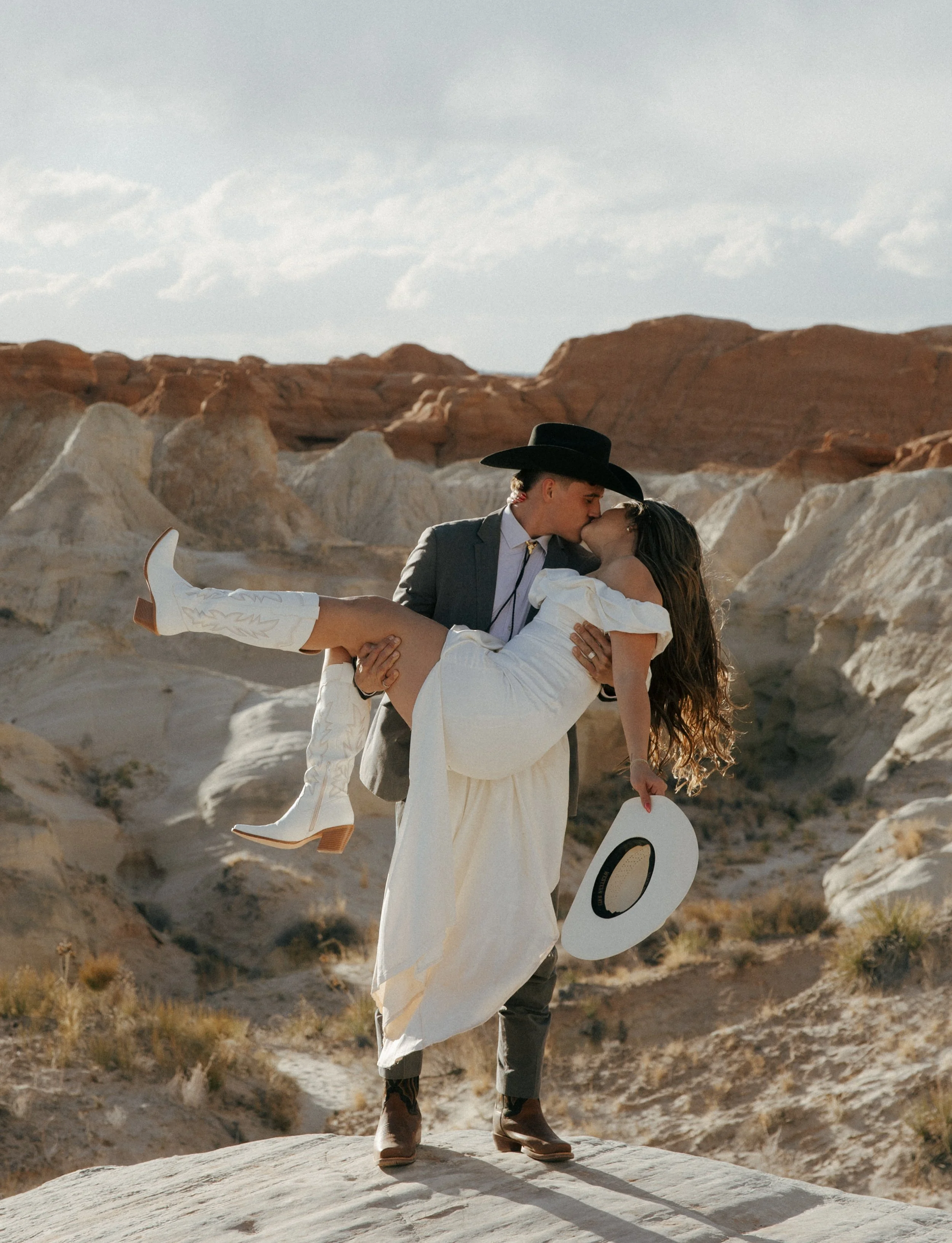 A couple dressed in Western-style clothing sharing a kiss in a desert landscape while the man carries the woman in his arms, holding a white hat.