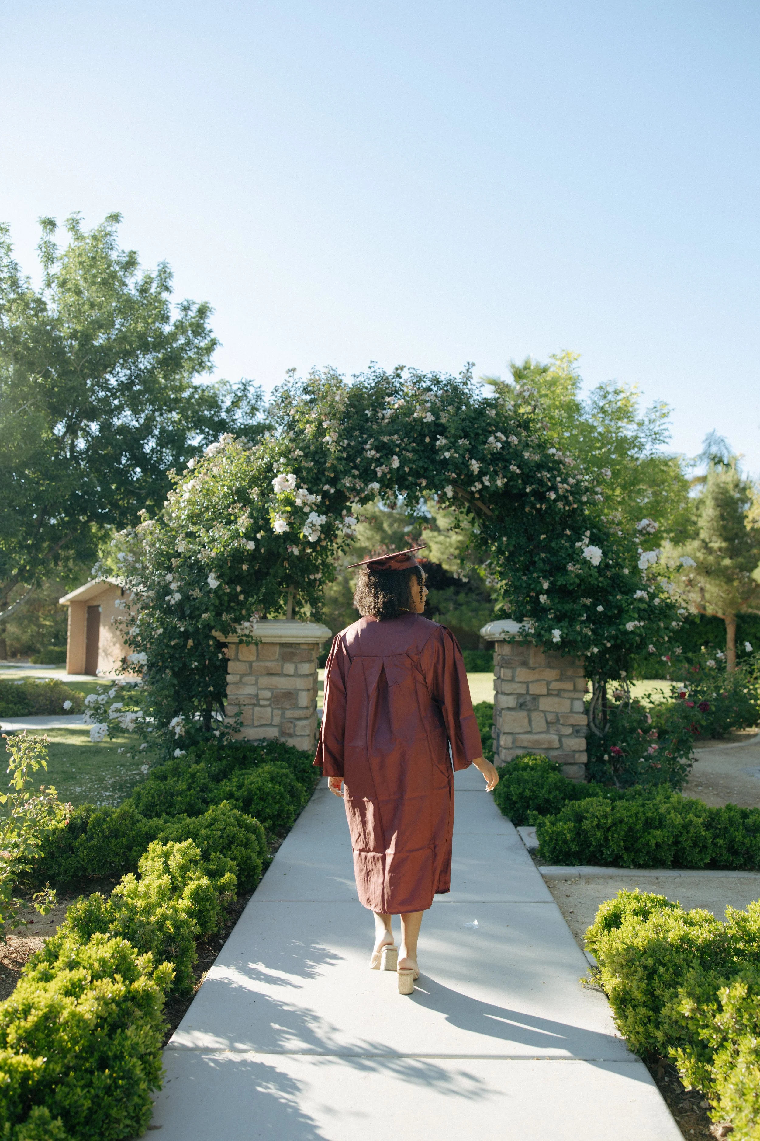 A woman in a maroon graduation gown and cap walking on a sidewalk through a garden archway with white flowers.