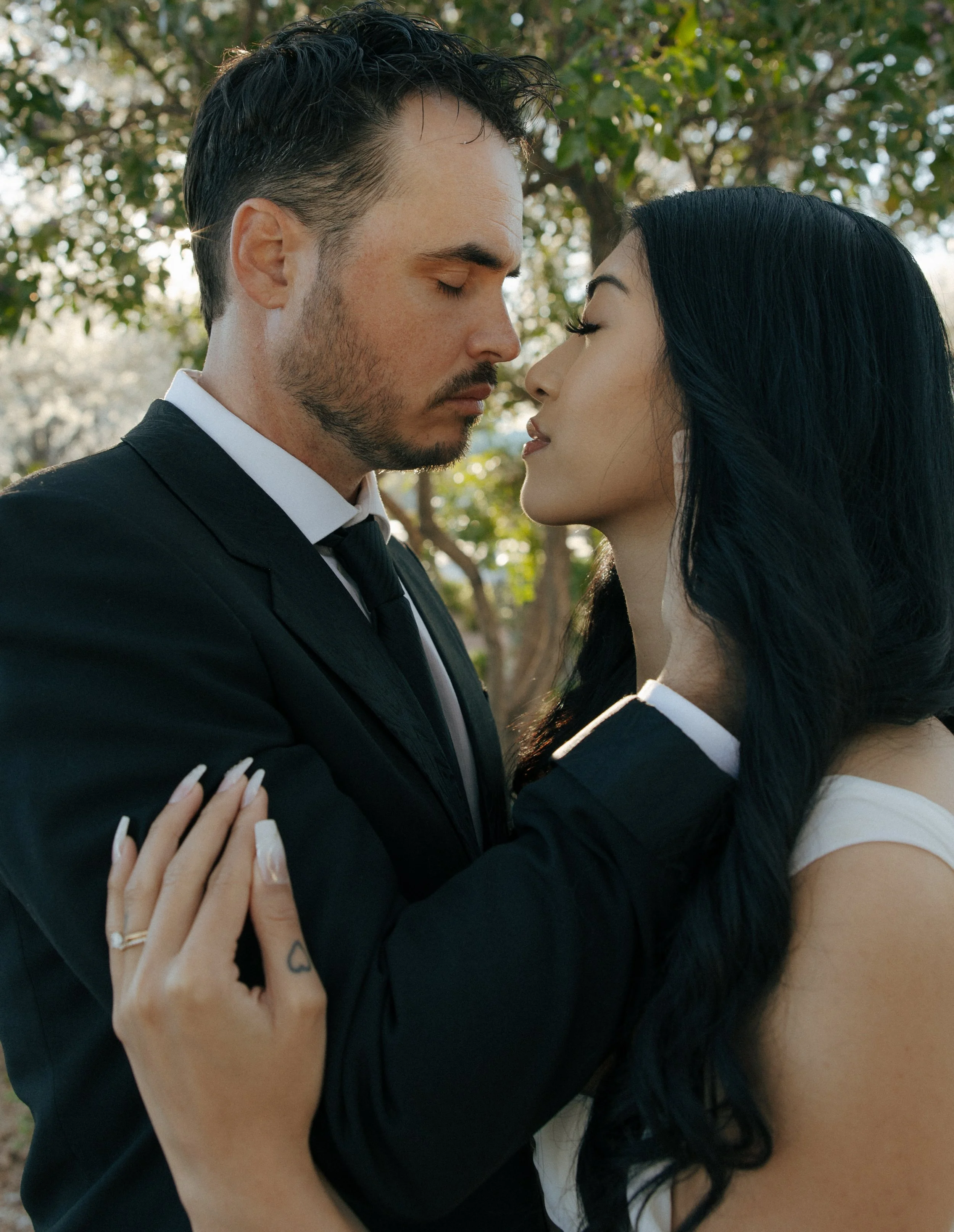 A man in a black suit and a woman in a white dress are standing close together outdoors with trees in the background, both with eyes closed.