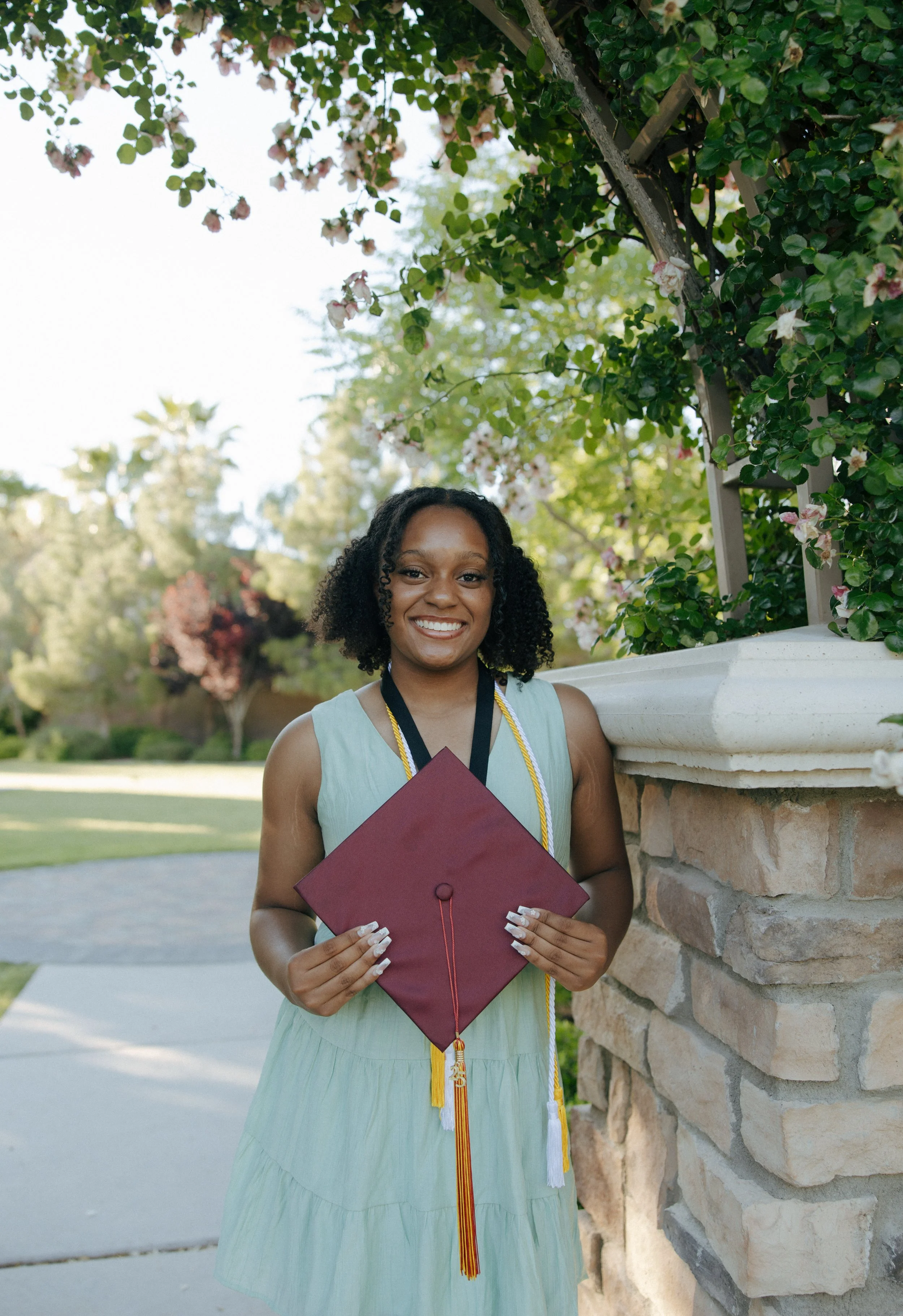 A young woman in a light green dress holding a maroon graduation cap outside, smiling, with flowering bushes and trees in the background.