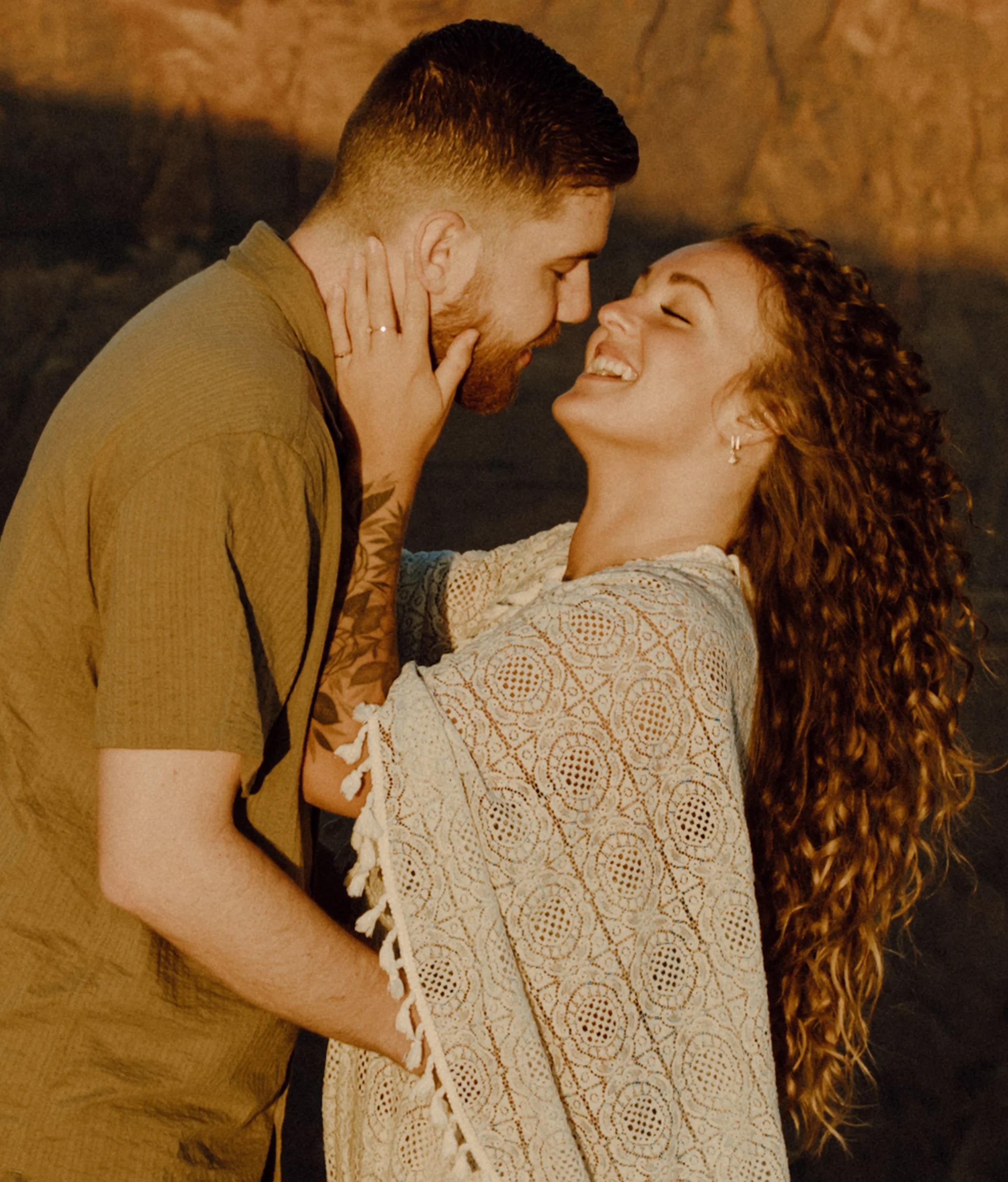 A man and woman are close together, smiling and gazing at each other affectionately. The man is holding the woman and she has her hand on his face, with both leaning in close.