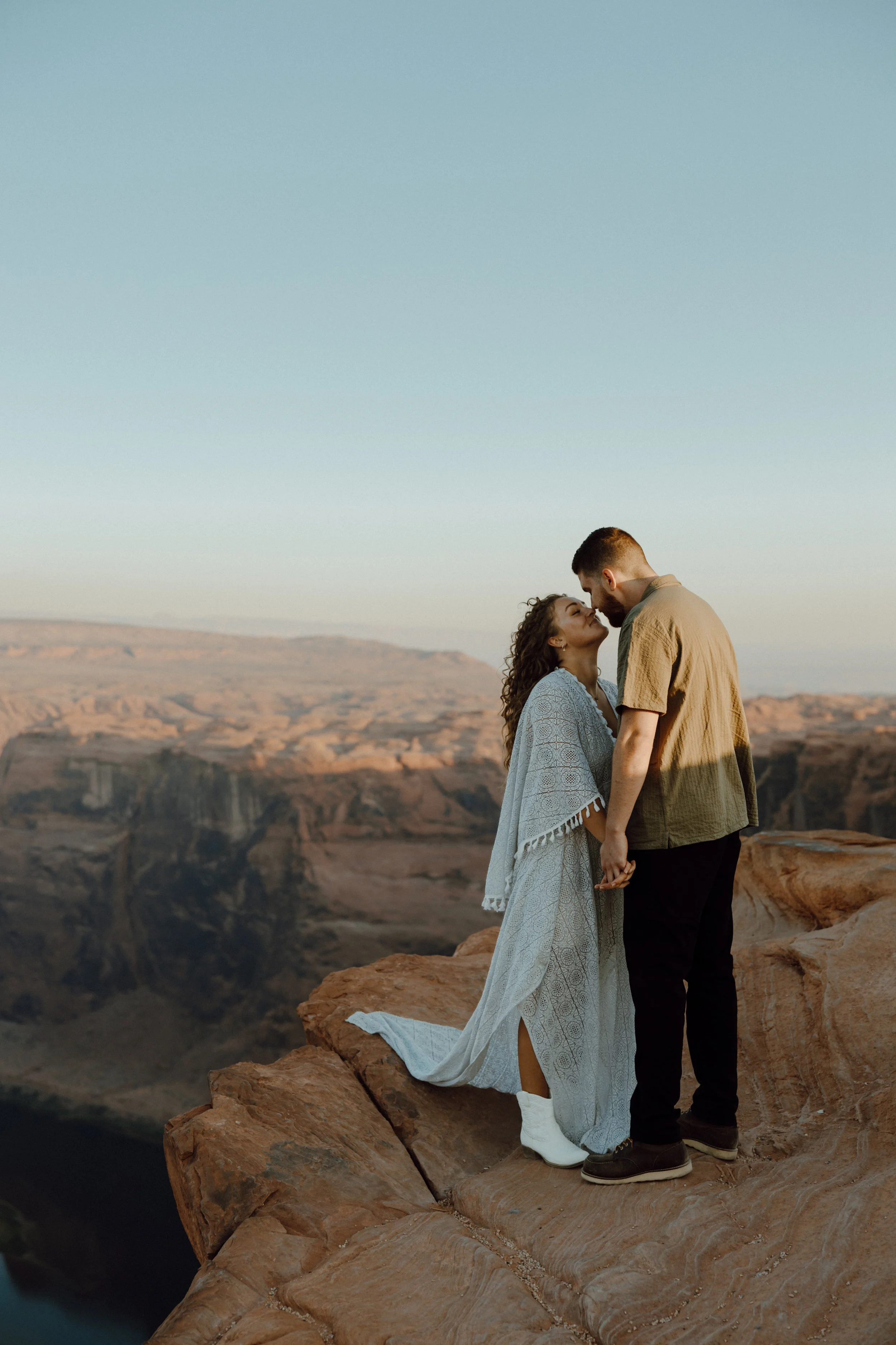 A couple standing on rocky cliffs holding hands, about to kiss with a canyon in the background during sunset or sunrise.
