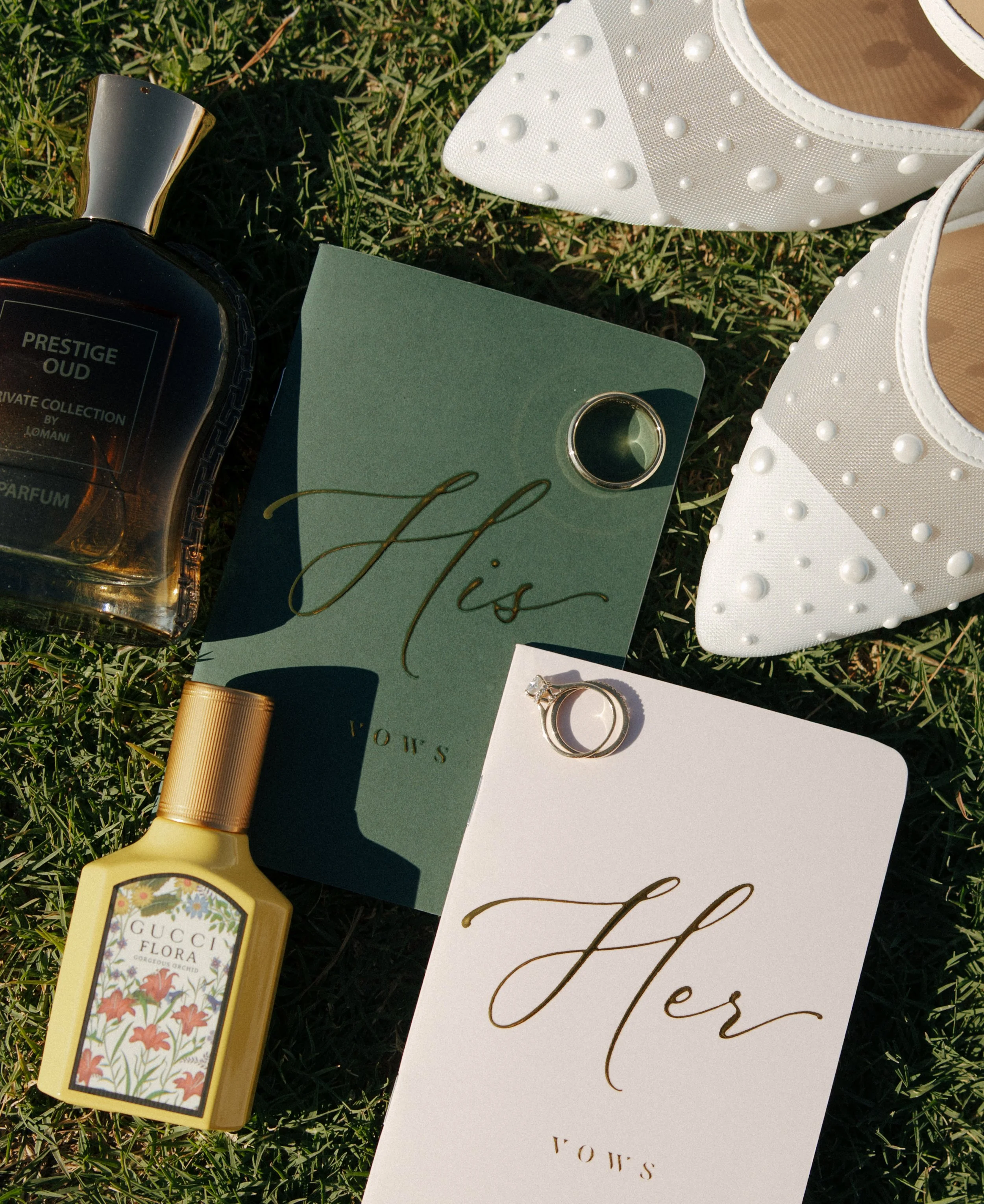 White embellished shoes, perfume bottles, herbal ring, and wedding rings on green grass, arranged in a romantic setting.
