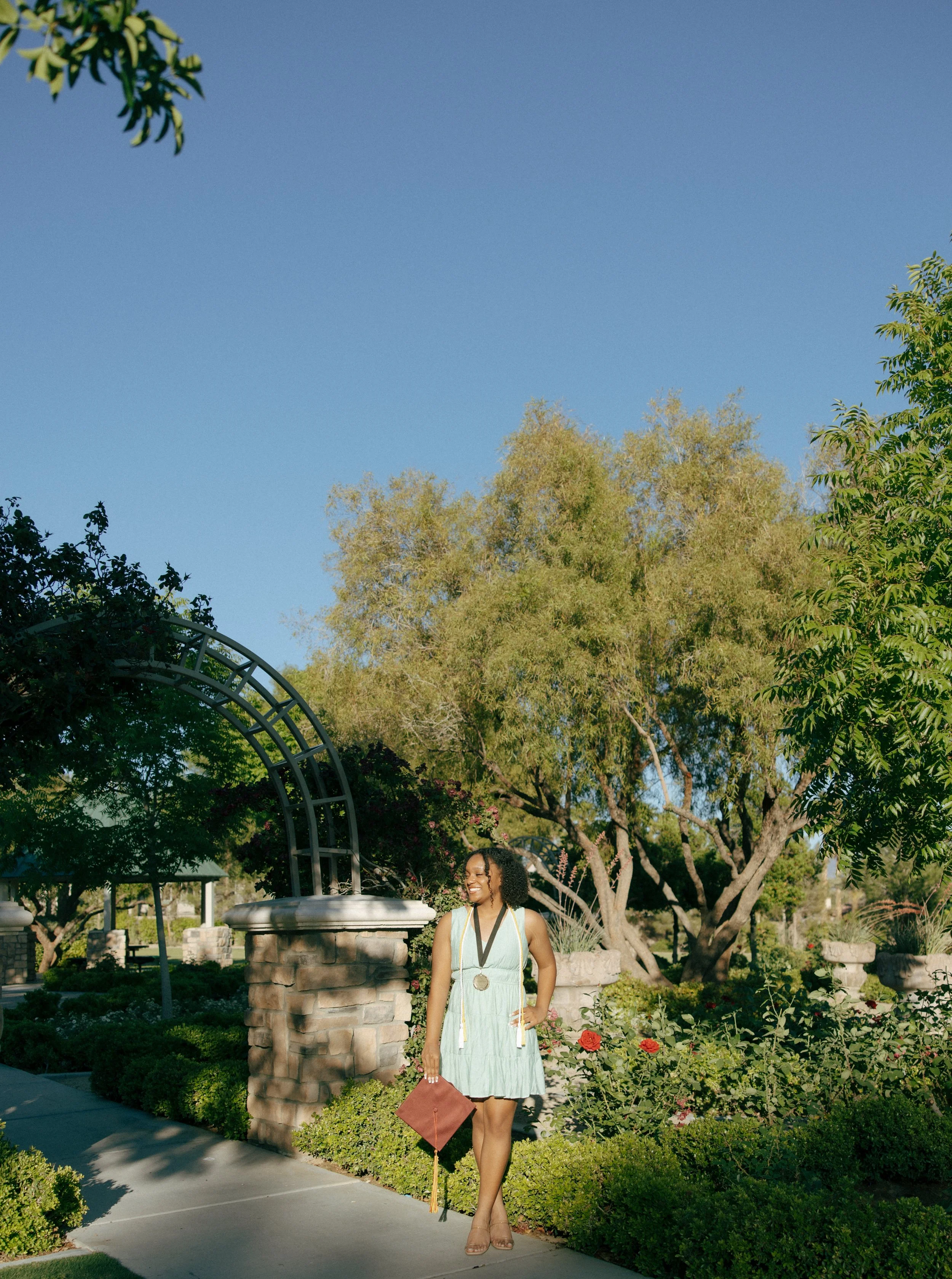 A woman in a light blue dress with a graduation medallion, holding a diploma, standing on a sidewalk in a park with trees, flowers, and a brick and metal archway, smiling.