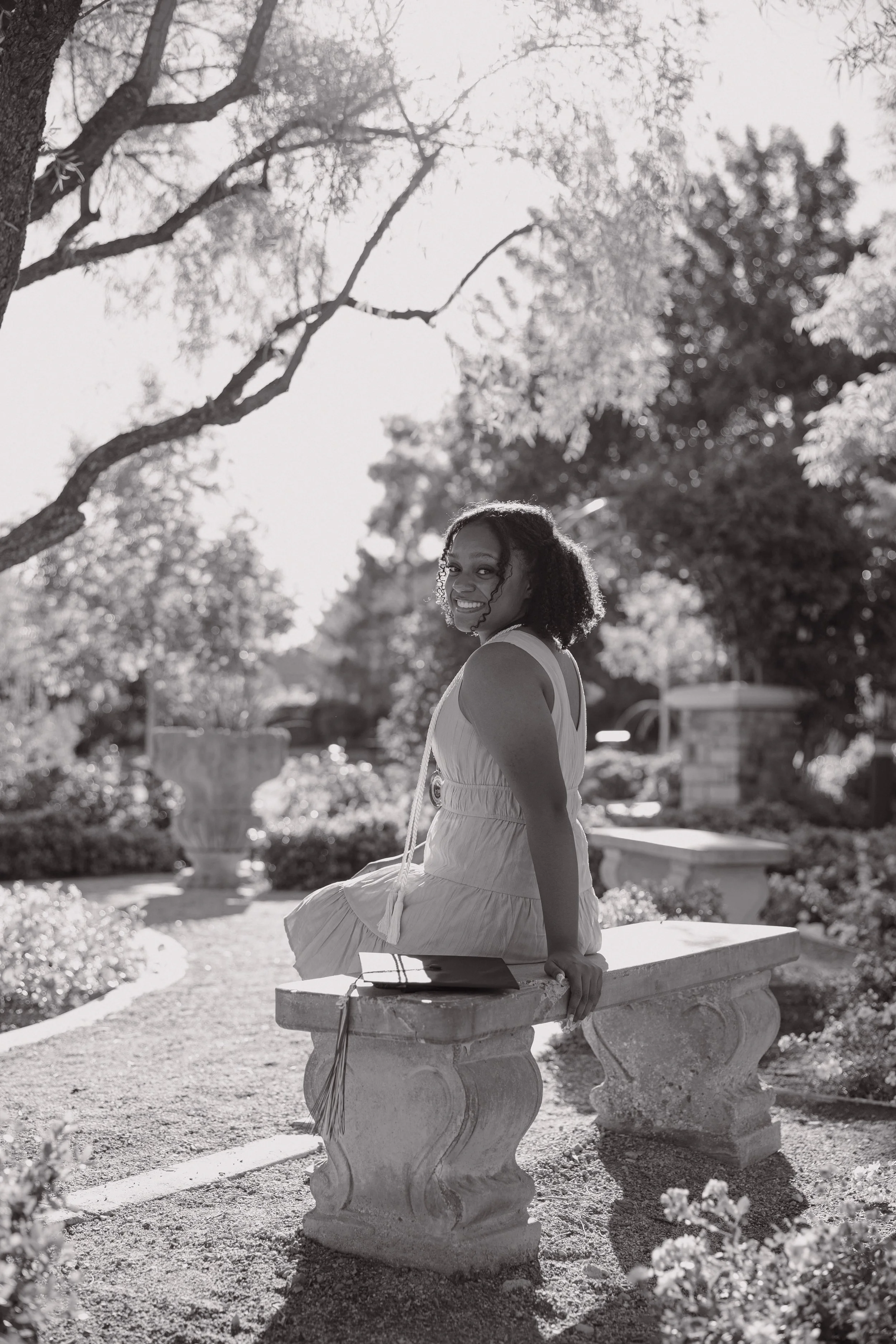 A young woman sitting on a stone bench outdoors in a park, smiling at the camera with trees and bushes in the background in black and white.