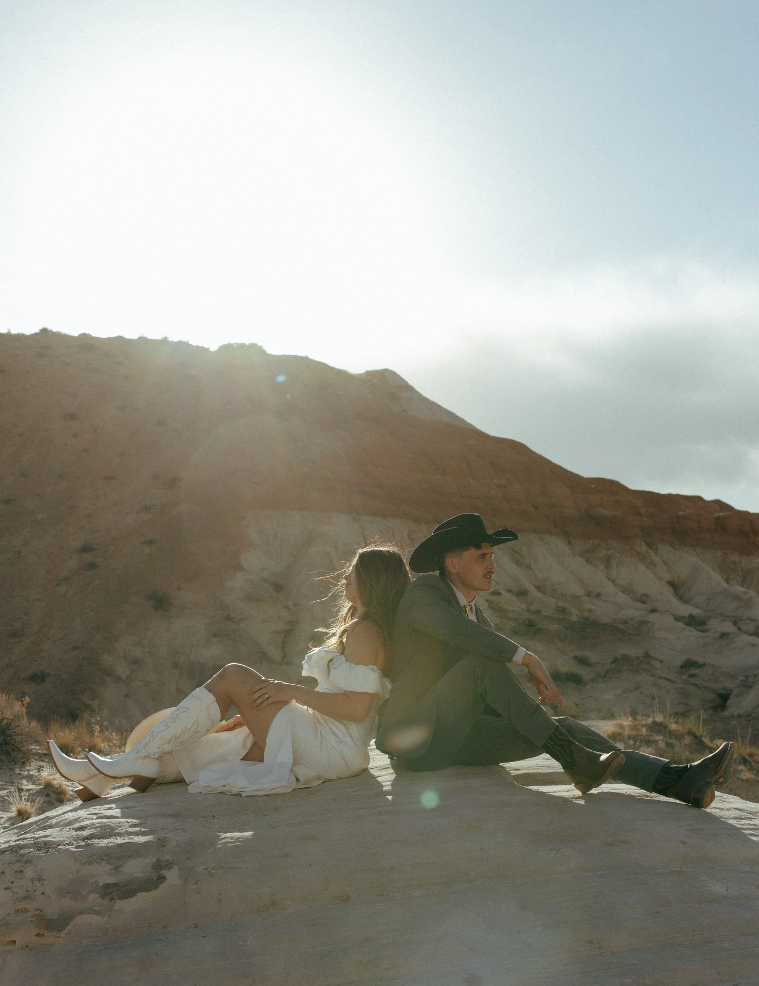 A man and woman sitting back-to-back on a rock in a desert landscape, with mountains and partly cloudy sky in the background, bathed in sunlight.