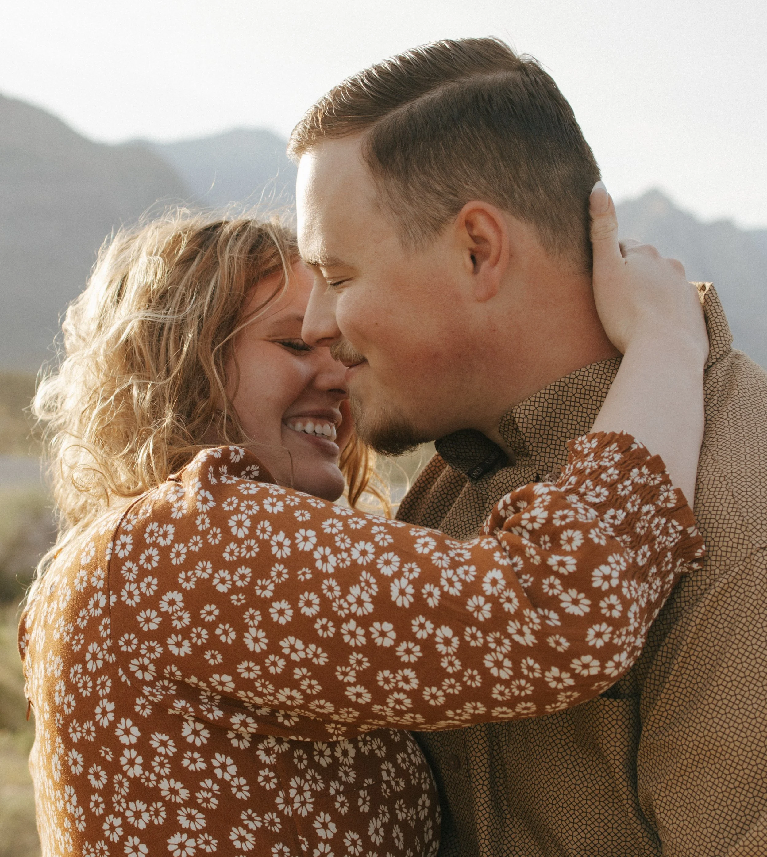A couple embracing outdoors with mountains in the background, smiling with their eyes closed.