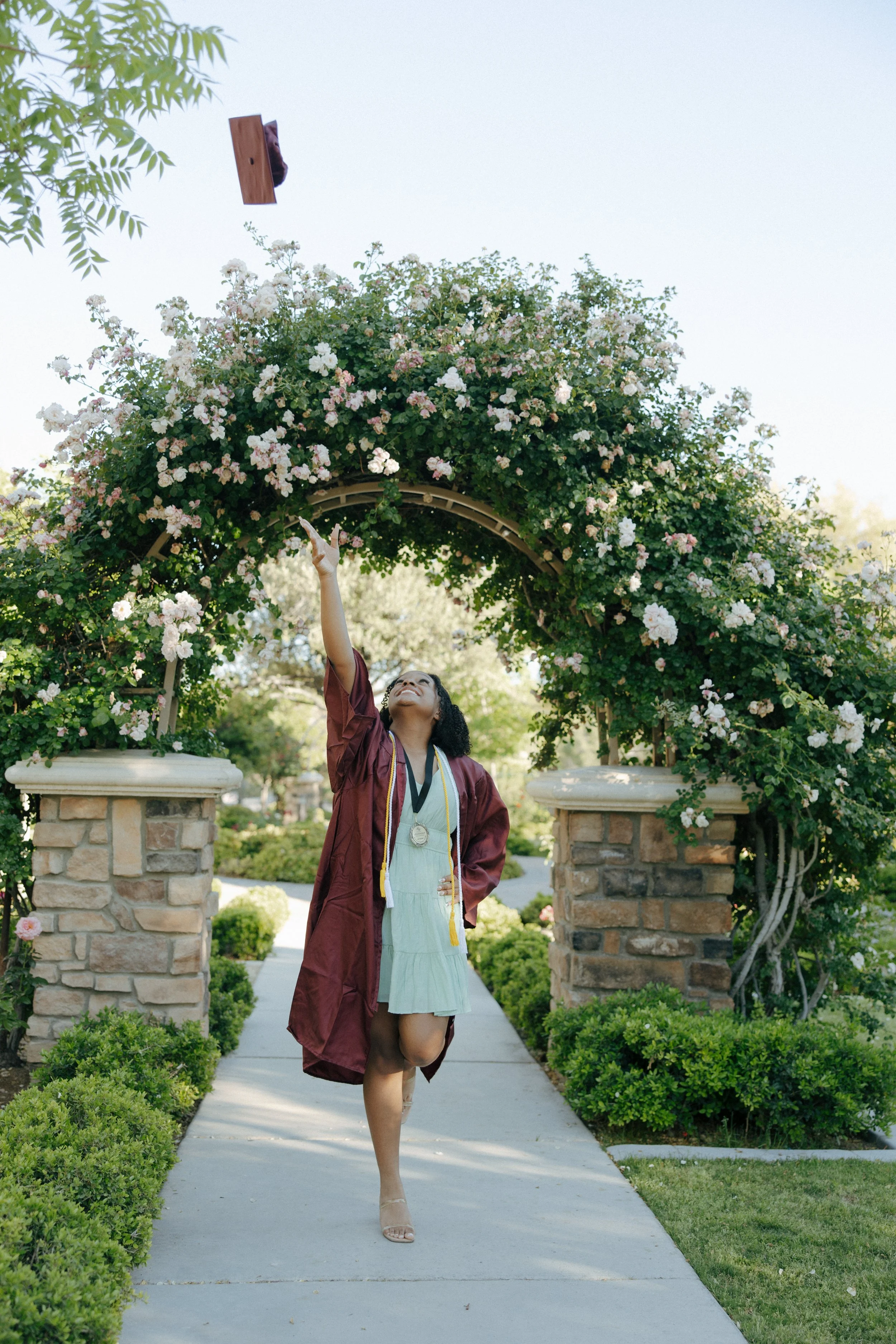 A woman in a graduation gown and cap throws her cap into the air while standing under a garden arch covered with pink and white flowers, celebrating her graduation.