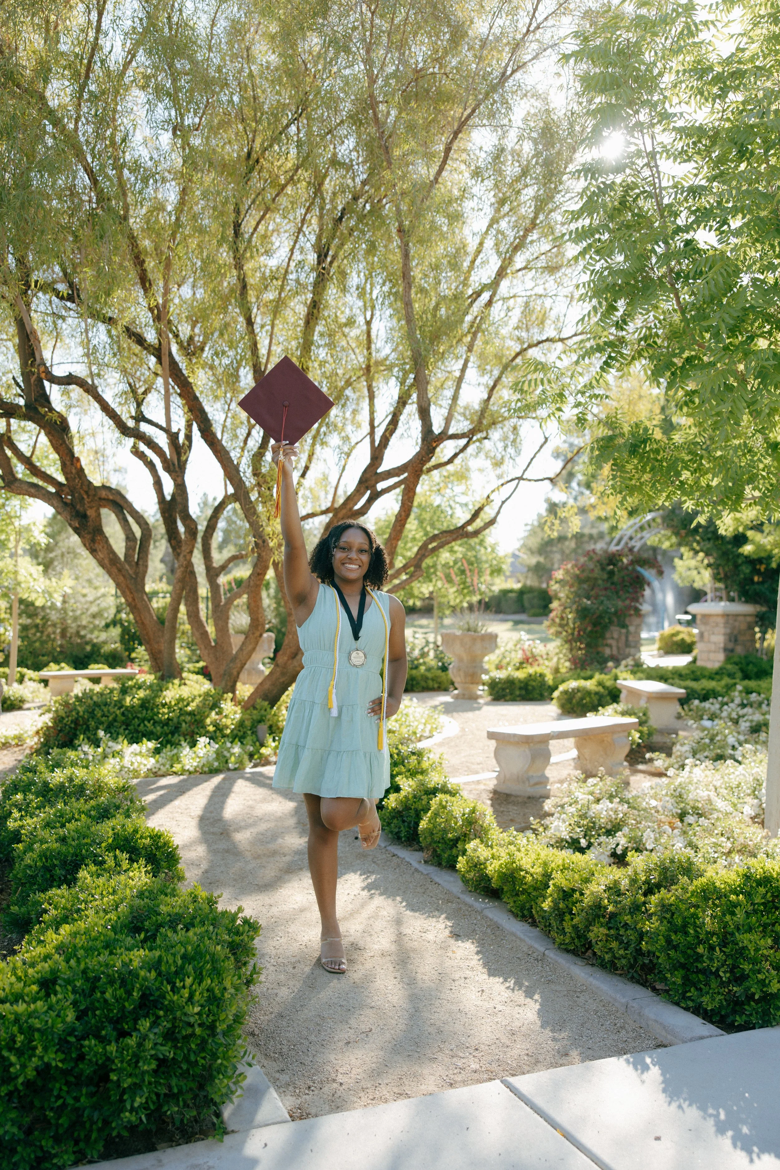 A woman celebrating her graduation outdoors in a sunlit garden, holding her cap in the air with a medal around her neck.