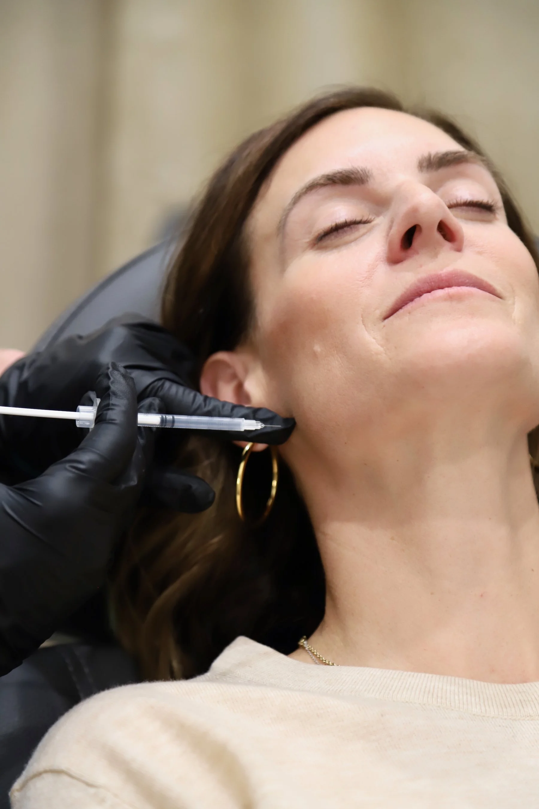 A woman with closed eyes receiving a cosmetic injection in her jaw area from a professional wearing black gloves.