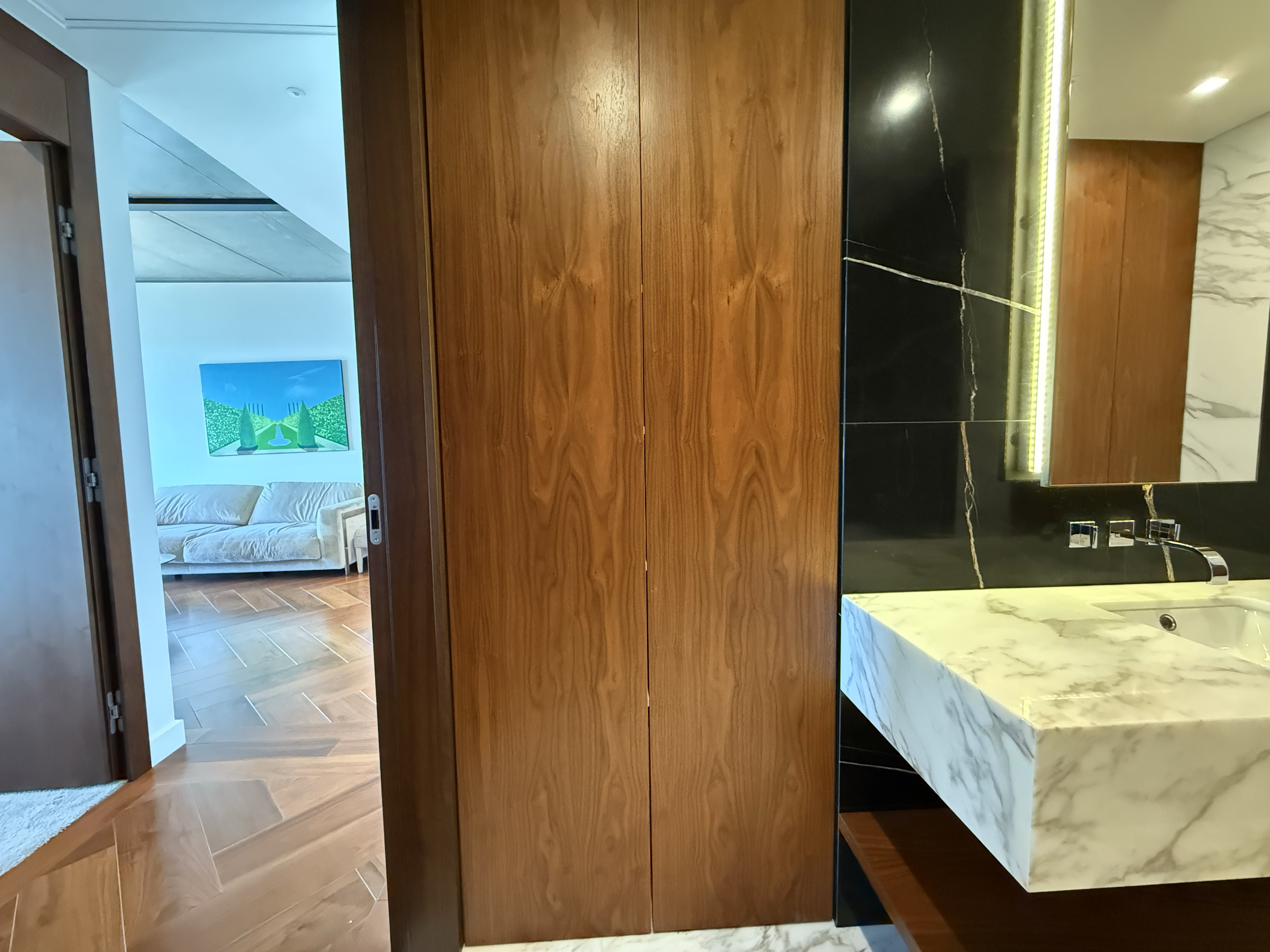 View of a modern bathroom with marble sink and black wall, wooden door and mirror, and a glimpse into a living room with a sofa and colorful artwork