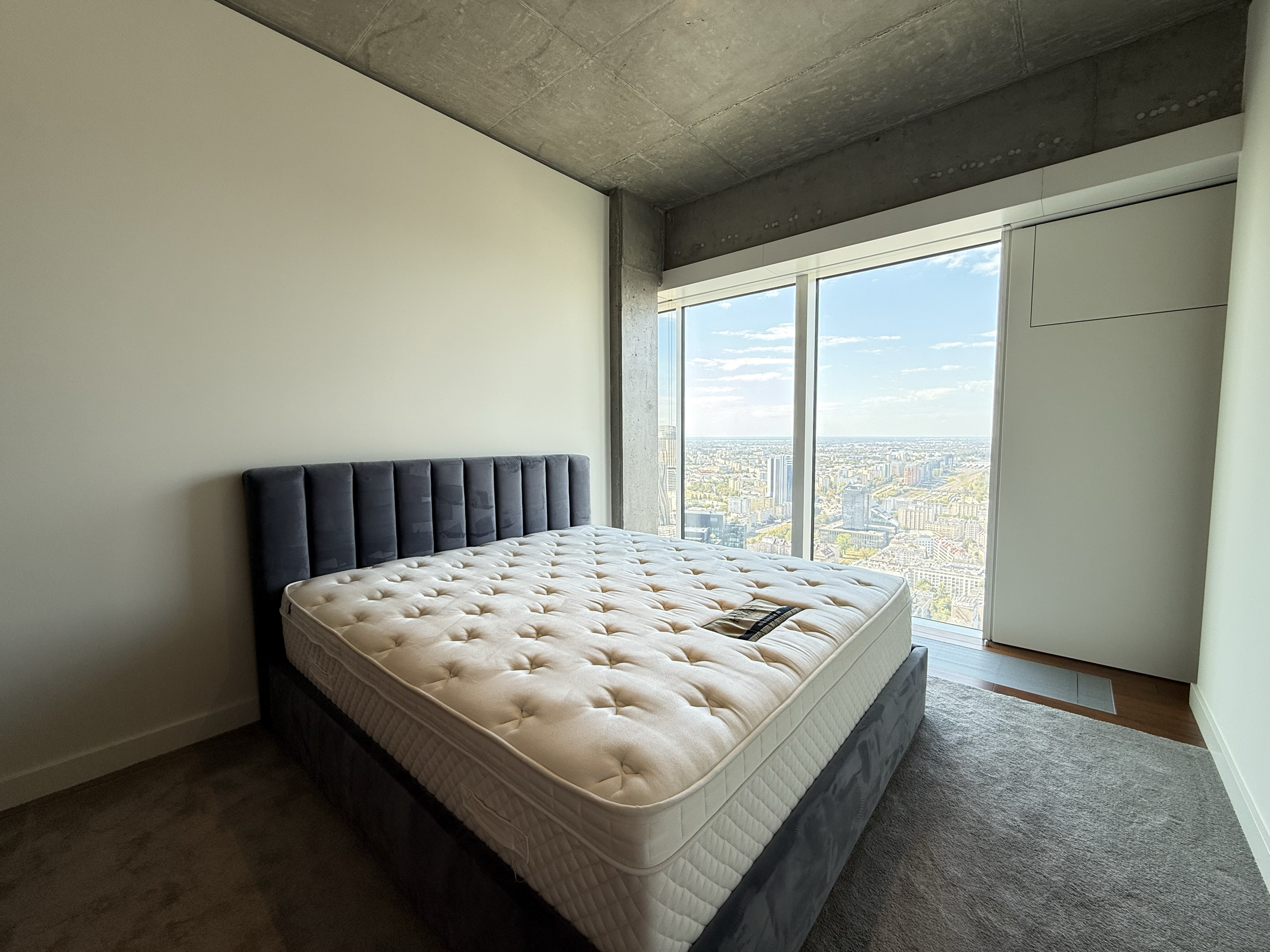 Empty bedroom in a high-rise apartment with a mattress on a bed frame, large window with a city view, concrete ceiling, and a white wall.