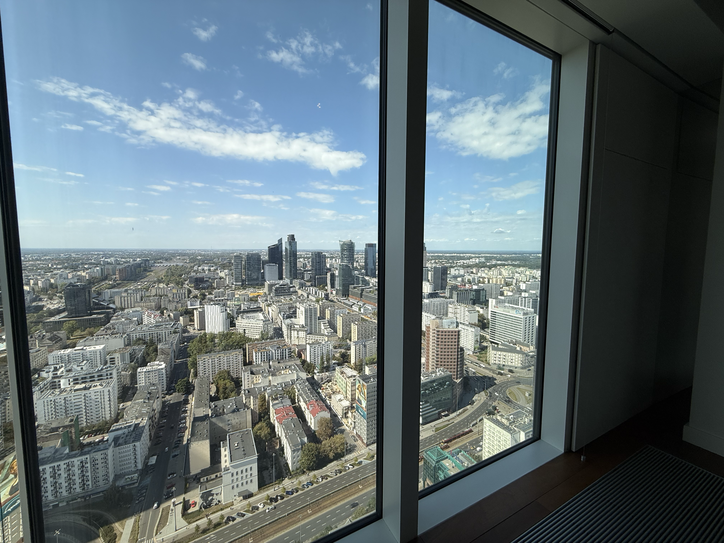 View of a city skyline through large floor-to-ceiling windows of a high-rise building with a clear blue sky and some scattered clouds.