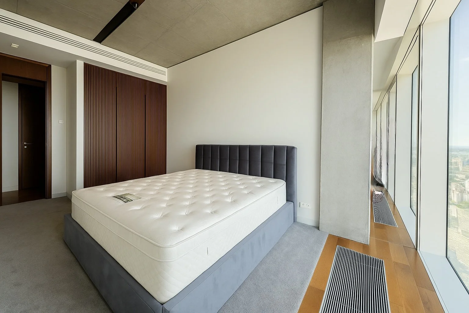 Modern bedroom with a large bed, upholstered headboard, wooden floors, and floor-to-ceiling windows showing a city view.
