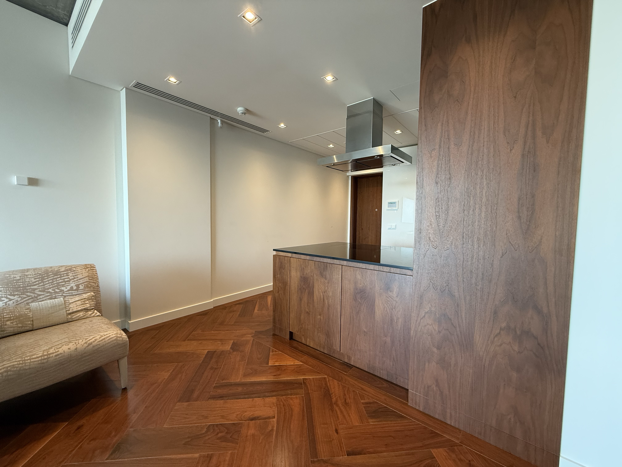 Empty modern kitchen with wooden cabinets, a stove, and a wooden accent wall, adjacent to a beige patterned sofa, with wood flooring and recessed ceiling lights.