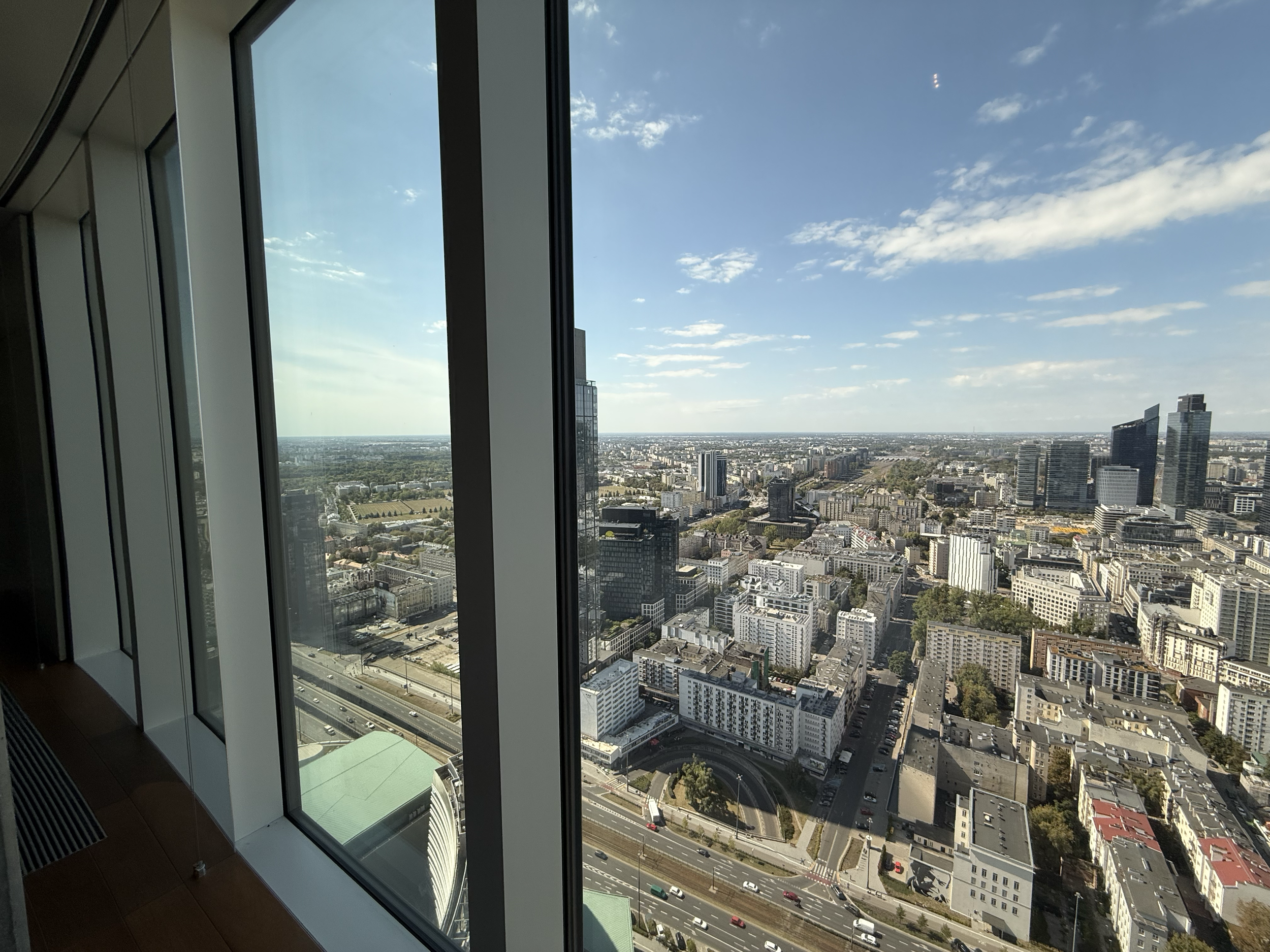 A view of a city skyline seen from a high-rise building window, showing various buildings, roads, and a partly cloudy sky.