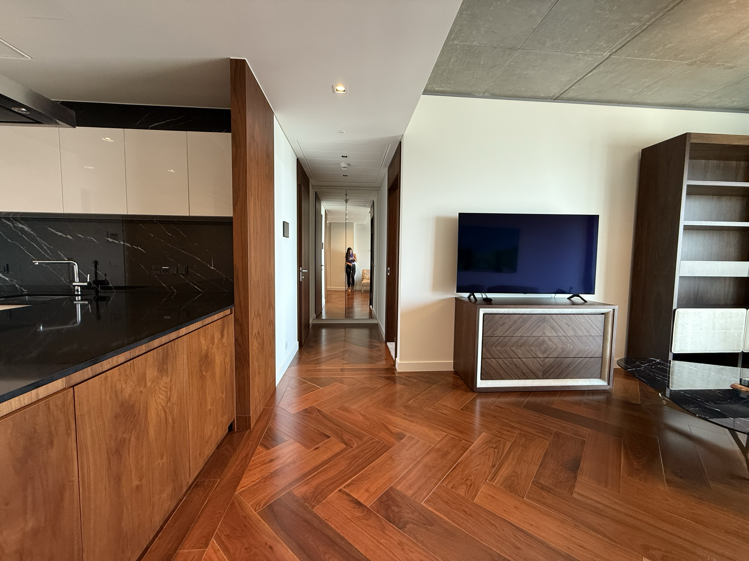 Living room with wooden floors, white wall, black TV on a wooden cabinet, and a kitchen counter with black marble surface on the left. A person is seen in the mirror at the end of the hallway.