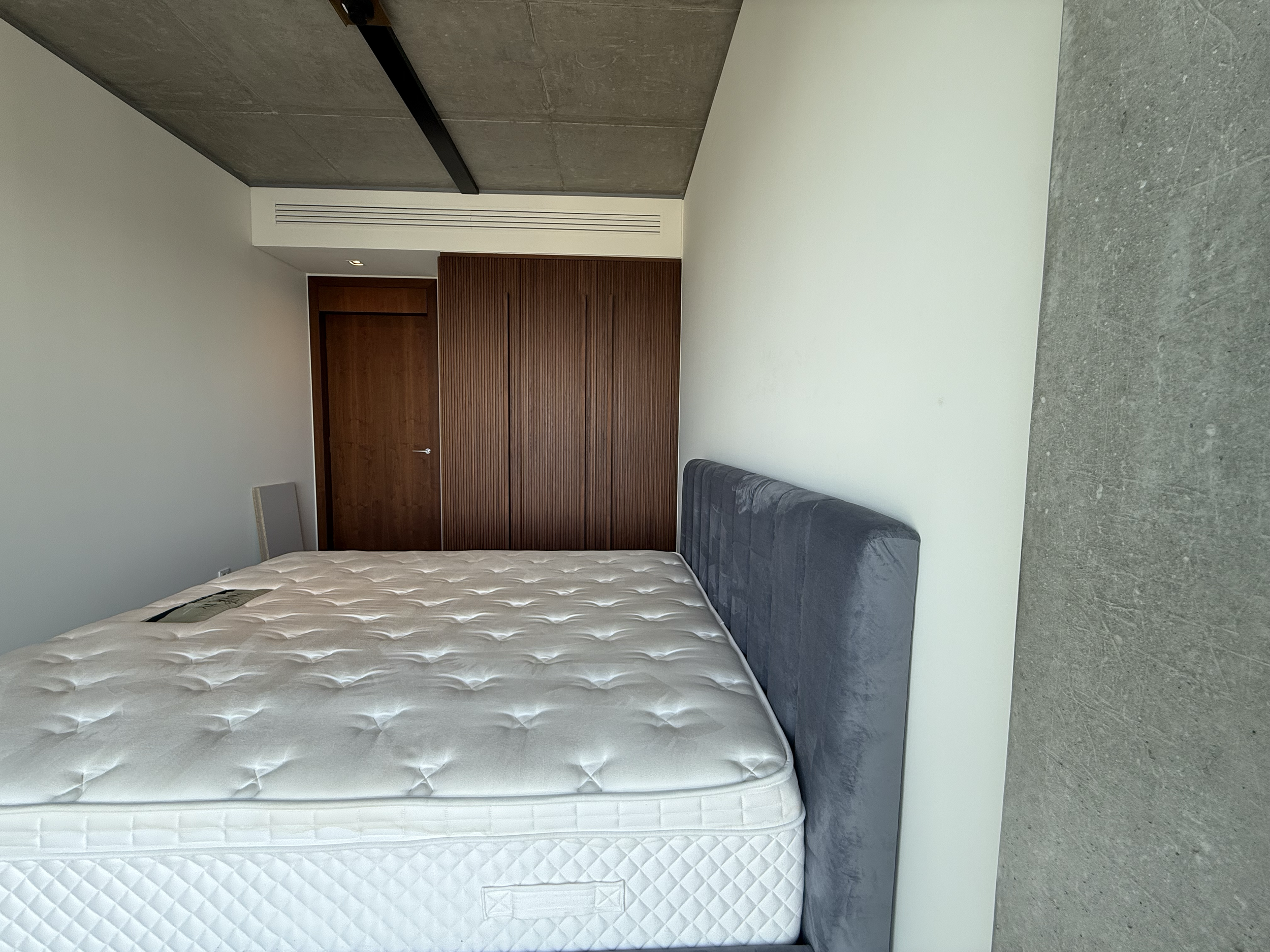 Modern bedroom with a large mattress, gray upholstered headboard, white walls, wood-paneled closet, concrete ceiling, and light gray concrete flooring.