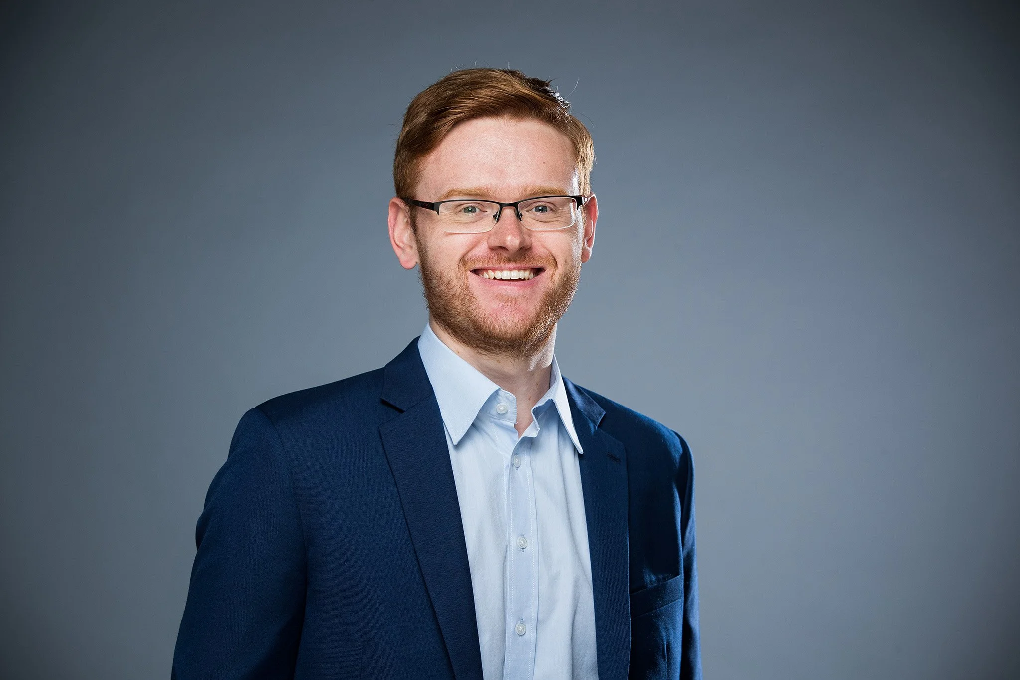 Ein lachender Mann mit Brille, rotem Haar und Bart, trägt einen blauen Anzug und ein weißes Hemd, vor einem grauen Hintergrund.