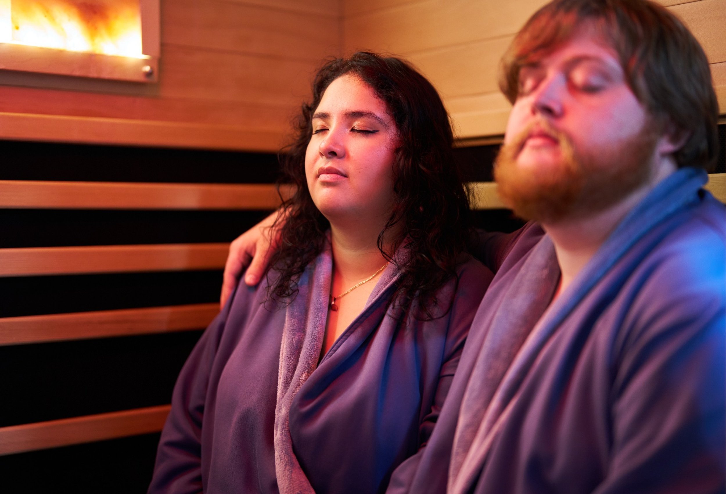 A woman and a man sitting against a wooden wall with their eyes closed, appearing relaxed and peaceful.