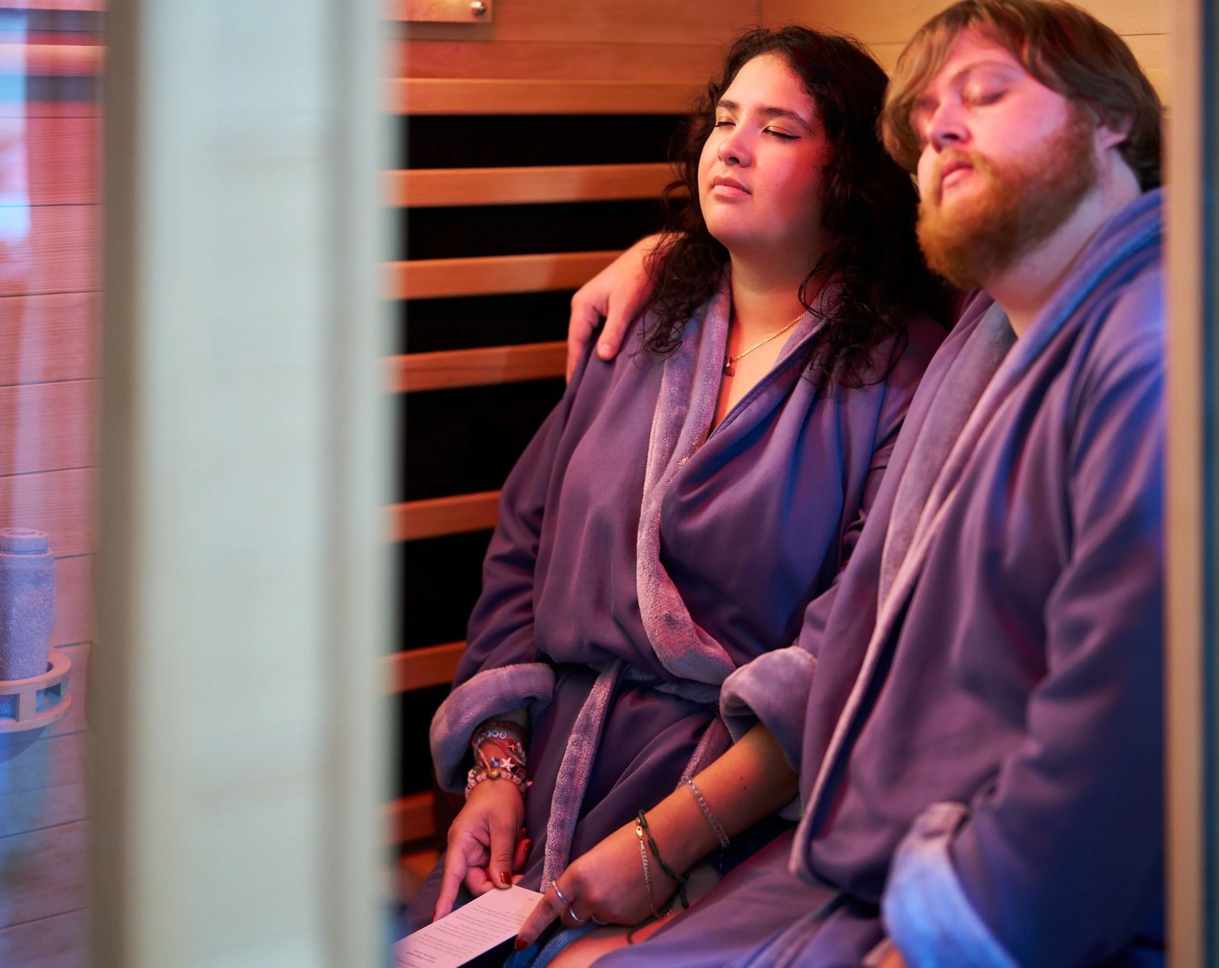 A woman and a man sitting closely together in a sauna, wearing robes and appearing relaxed with their eyes closed.