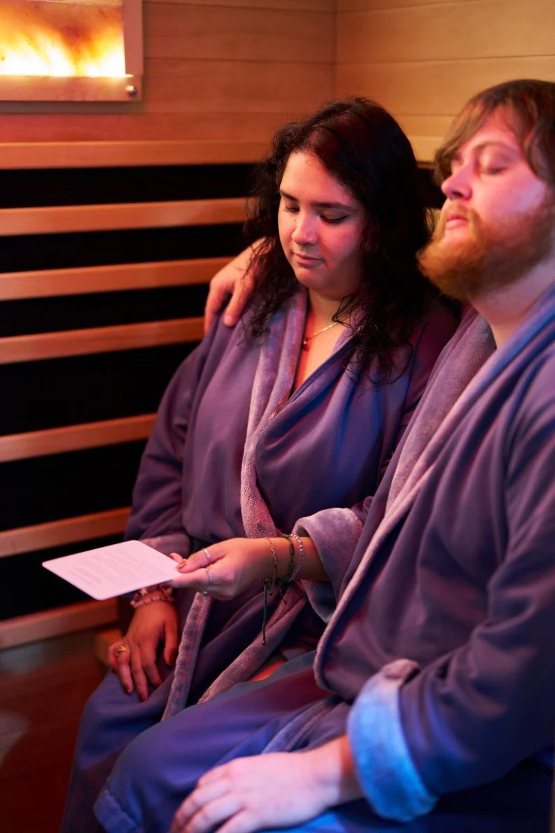 A woman and man sitting close together in a cozy, dimly lit room, wearing robes, with the woman holding a piece of paper.