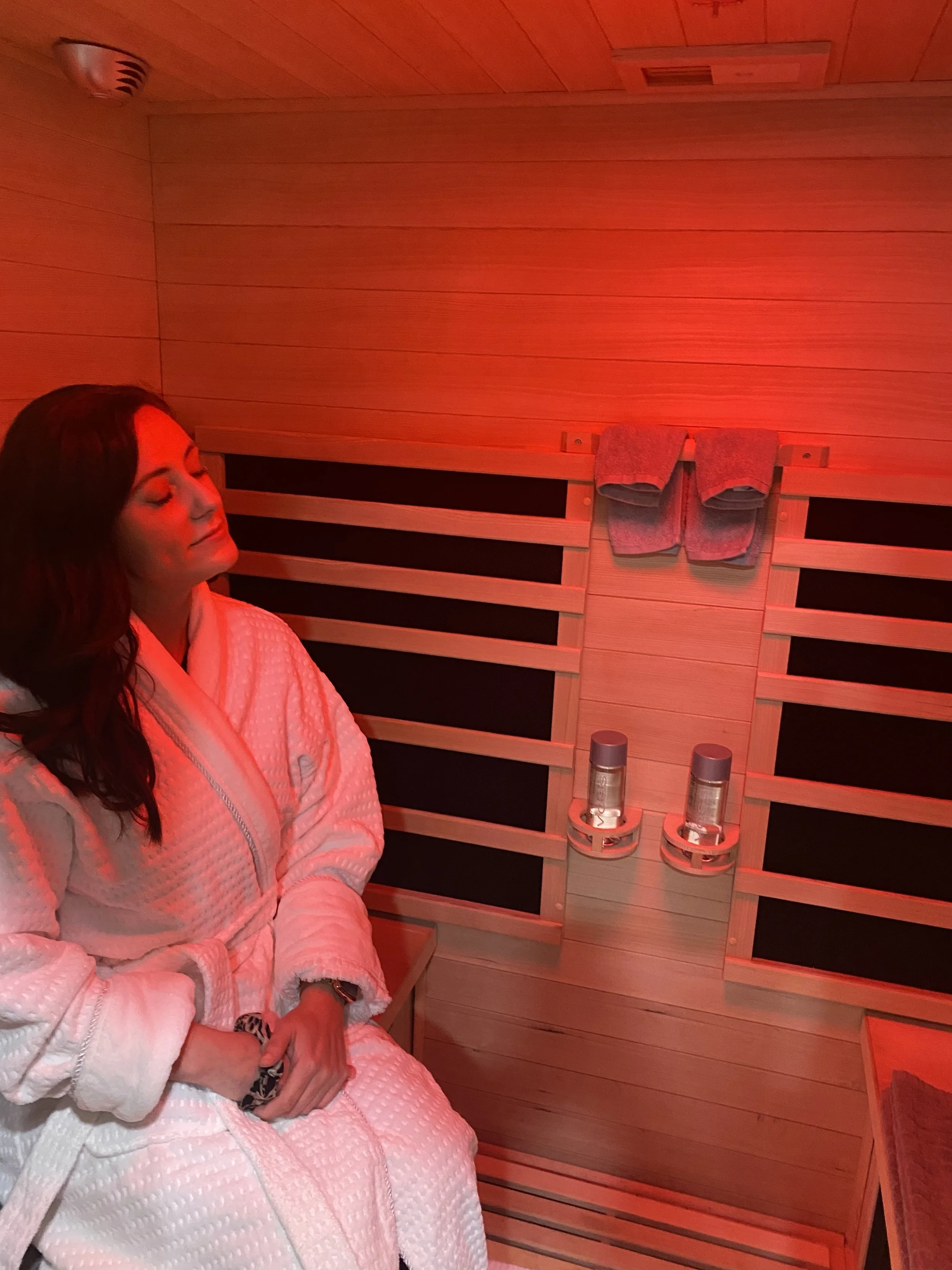 Woman relaxing inside a sauna, wearing a white bathrobe, with a peaceful expression, surrounded by wooden walls and red lighting.