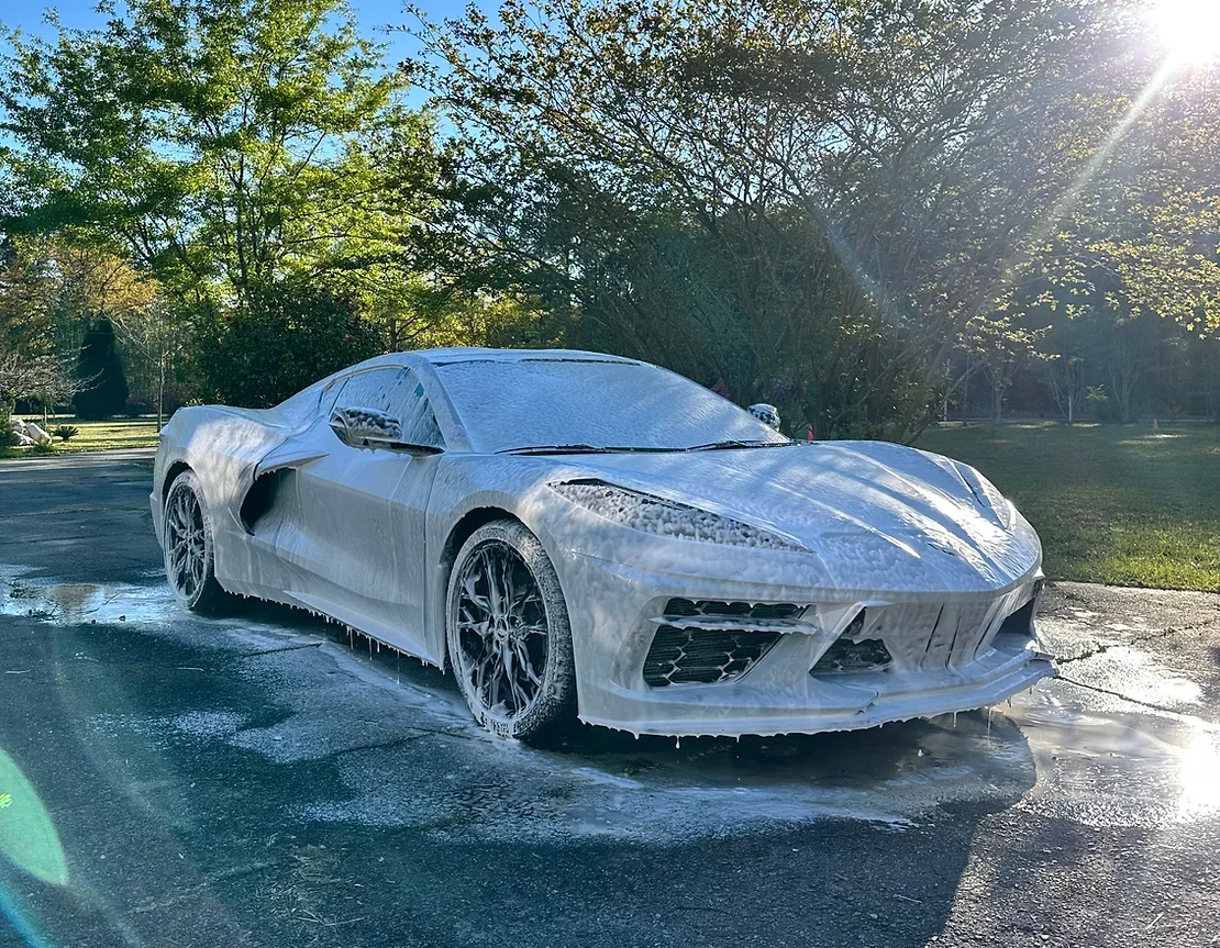 A white sports car covered in soap suds during a wash, with foam dripping from its surface, parked on a wet driveway in a sunny outdoor setting surrounded by trees.