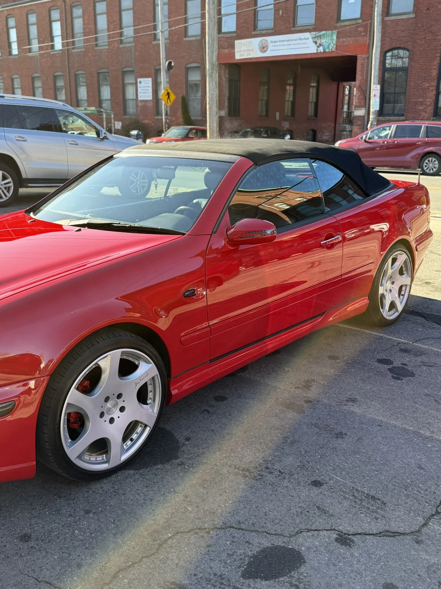 Red convertible car with black soft top parked on the street near a brick building, with other cars visible in the background.