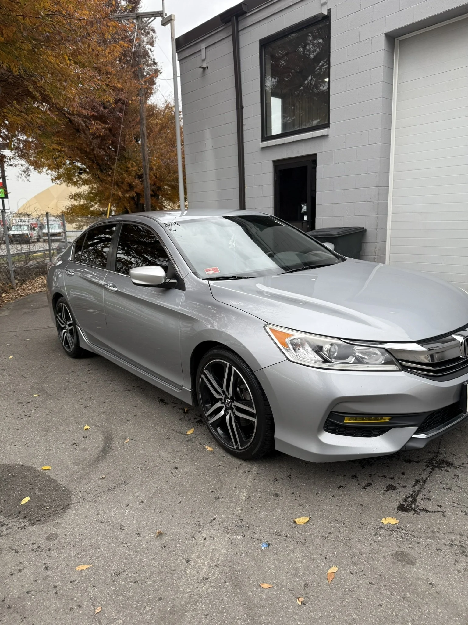 A silver sedan car parked outdoors on pavement near a gray building with a black door and window. Autumn trees with orange and brown leaves are visible in the background.